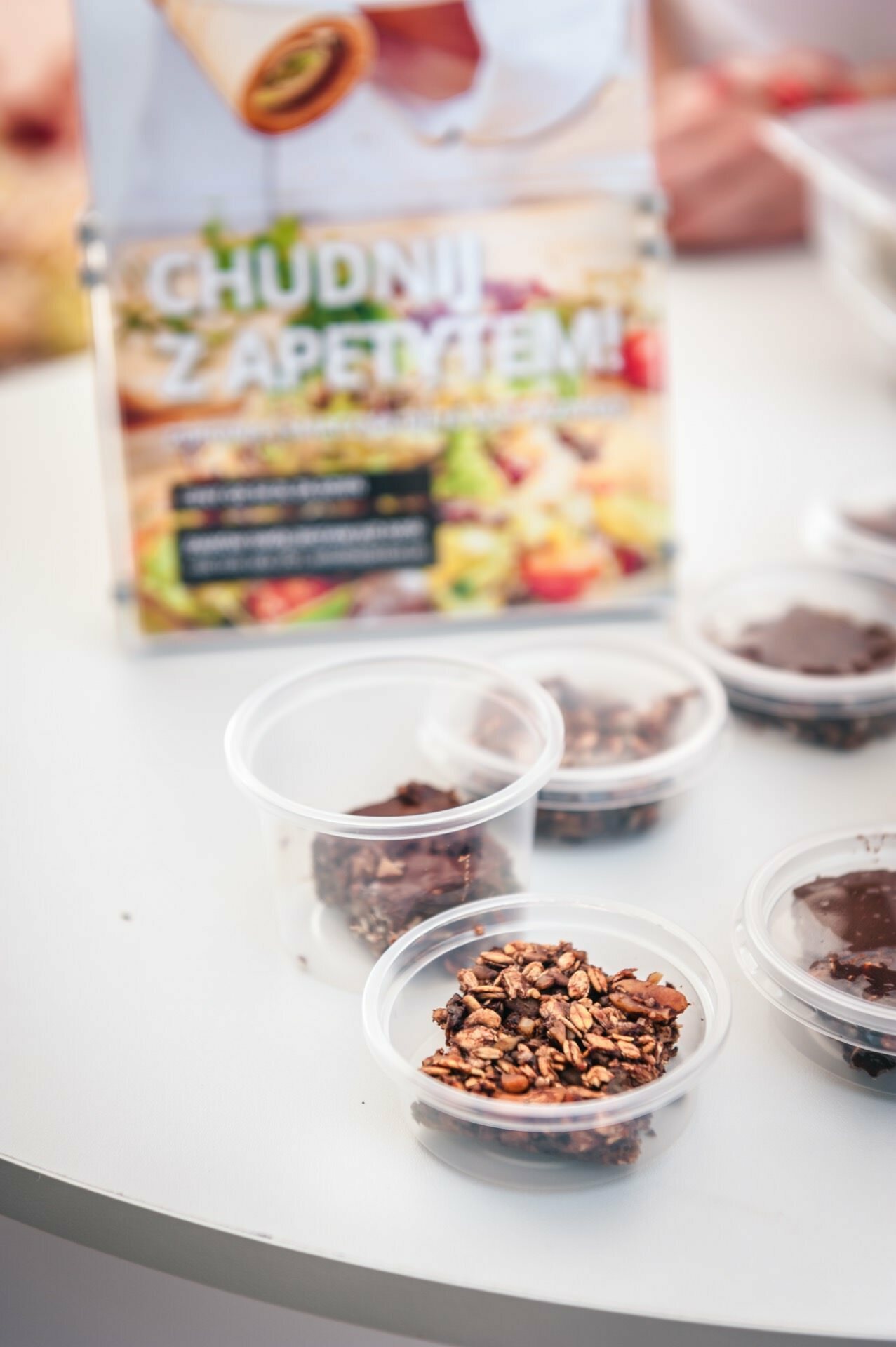 Small plastic cups containing muesli and chocolate are stacked on a white table. In the background, a signboard with a partially visible advertisement in Polish captures the essence of our photo report from the fair. 