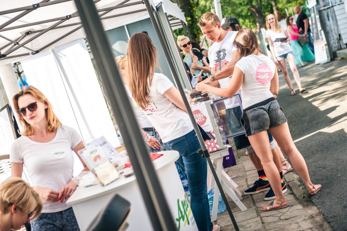 Ludzie wchodzą w interakcje przy stoisku na świeżym powietrzu pod namiotem na targu lub rynku. Wystawiane są różne produkty, a osoby są swobodnie ubrane. Kilka osób w tle cieszy się słonecznym dniem, uwieczniając chwile na fotorelację z targów.