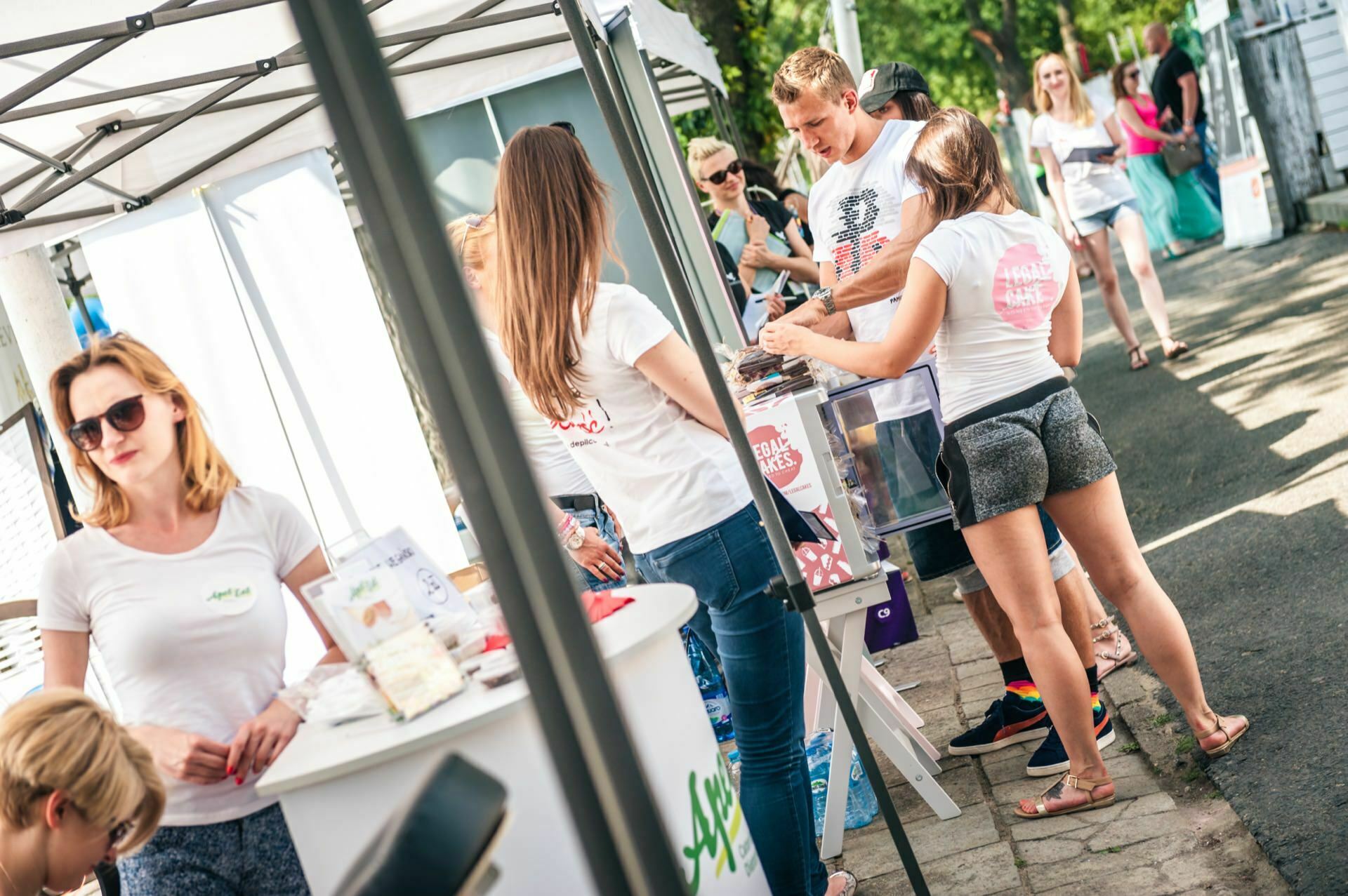 People interact at an outdoor booth under a tent in a market or marketplace. Various products are on display and people are dressed casually. A few people in the background are enjoying the sunny day, capturing moments for a photo essay of the fair.  