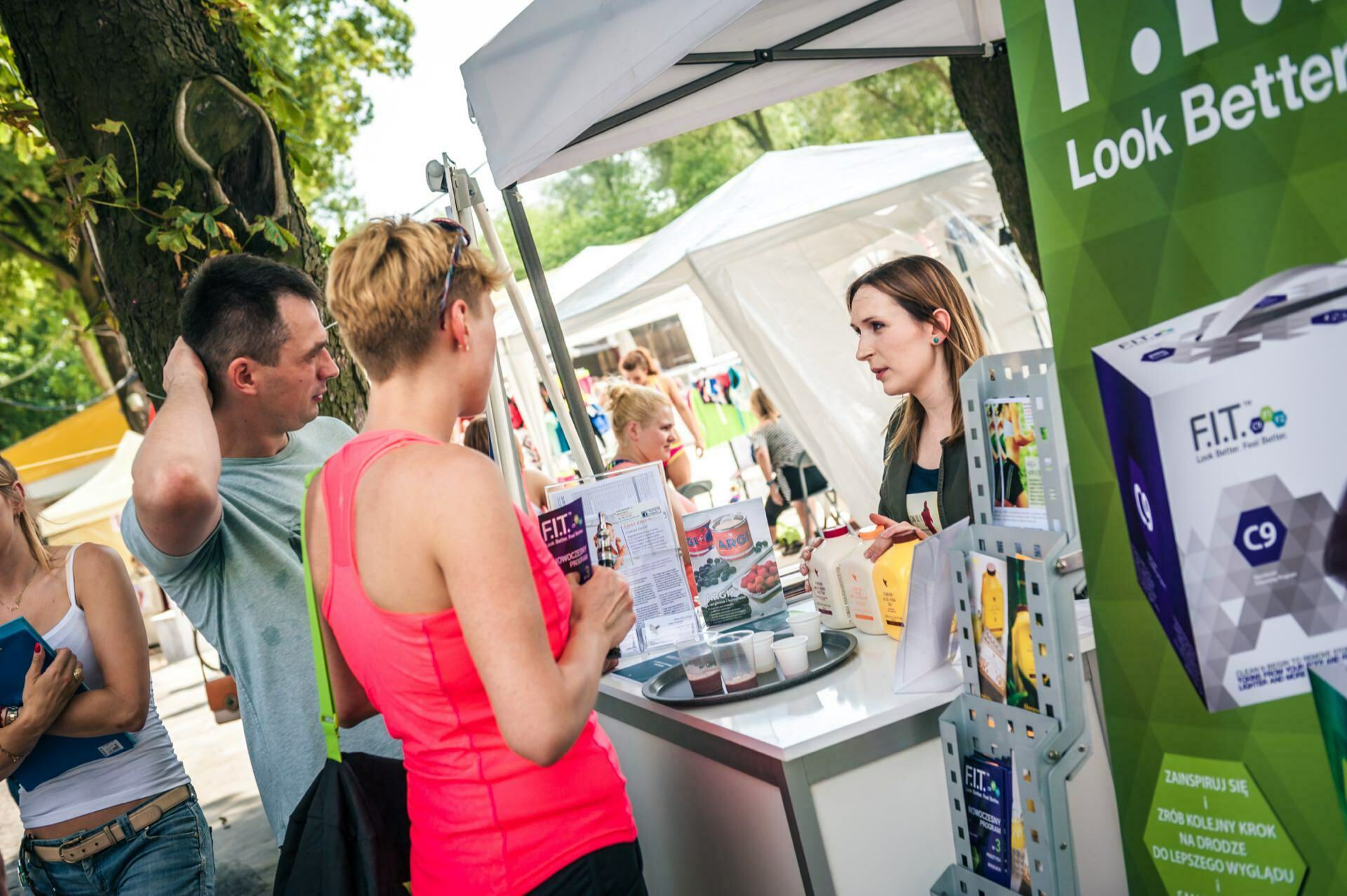 A photo report from the fair shows people at an outdoor booth interacting with a vendor. A woman in a pink T-shirt is reading a brochure, while the man next to her listens intently. The booth displays products related to health and well-being, including a large sign and boxes with "F.I.T." branding.  