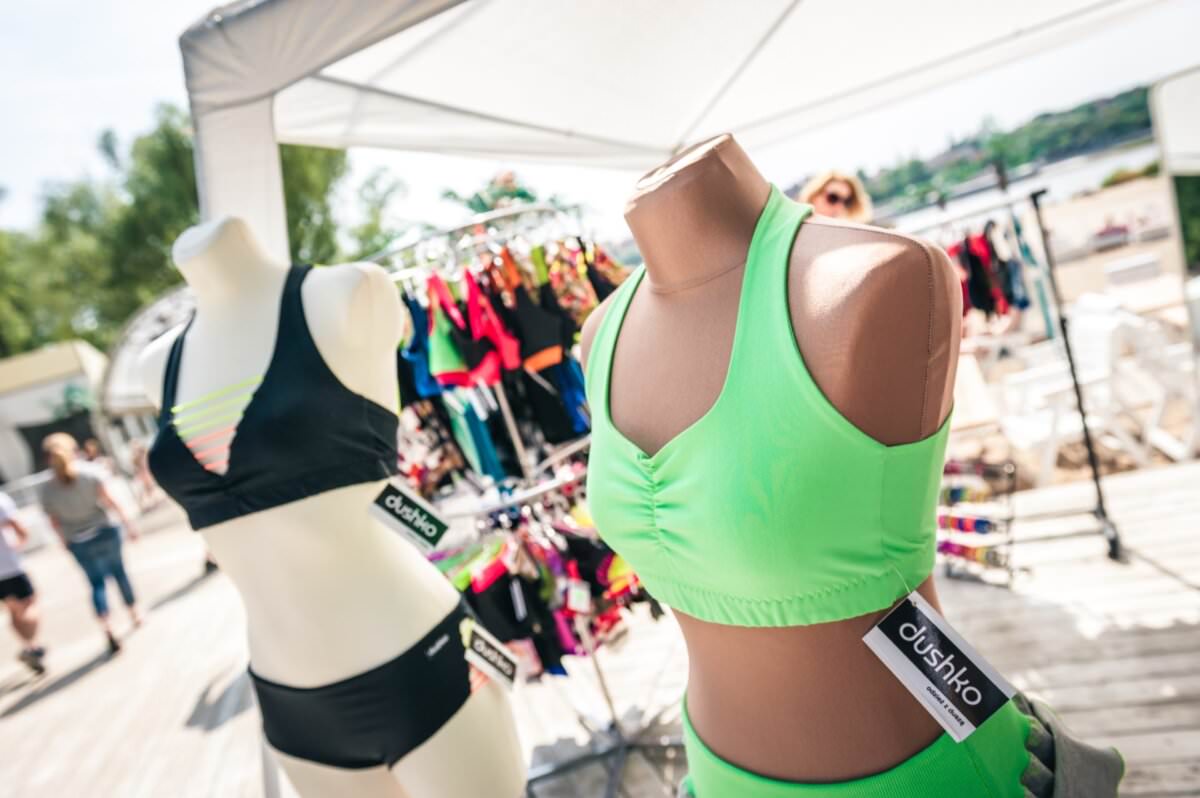 Dwa torsy manekinów eksponują stroje kąpielowe pod baldachimem na targu na świeżym powietrzu. Lewy, biały manekin nosi czarne bikini, podczas gdy prawy, brązowy manekin prezentuje jasnozielony dwuczęściowy zestaw. Różnorodne inne kolorowe stroje kąpielowe wiszą na wieszakach w tle, oddając istotę fotorelacji z targów.