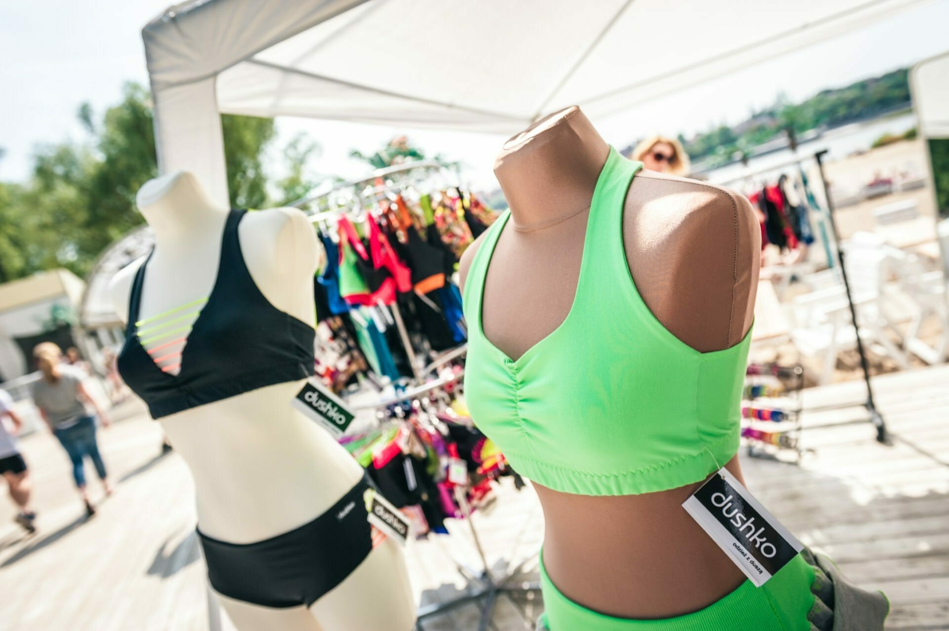 Two mannequin torsos display swimwear under a canopy at an outdoor market. The left, white mannequin wears a black bikini, while the right, brown mannequin displays a light green two-piece set. A variety of other colorful swimwear hangs on hangers in the background, capturing the essence of the photo fair.  