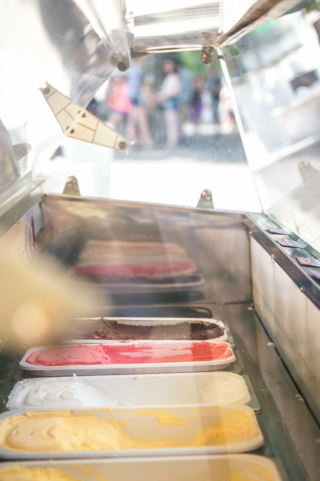 The image, reminiscent of a photo report from a trade fair, shows a view of the inside of an ice cream display freezer filled with ice cream of various flavors in tubes, visible through a transparent glass cover. Focusing on the ice cream, the blurry background shows people walking outside. 