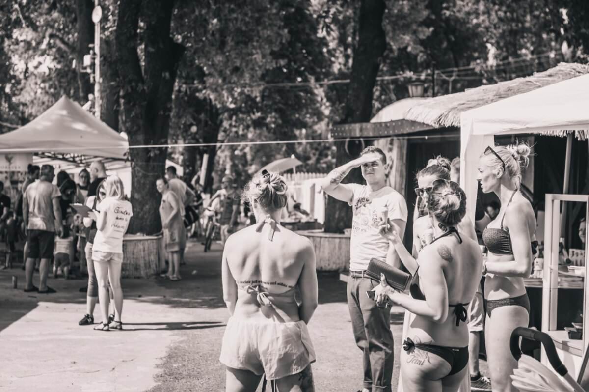 Grupa ludzi zbiera się na świeżym powietrzu, ubrana w stroje codzienne i plażowe. Niektórzy rozmawiają, inni stoją lub spacerują w tle. Scena obejmuje drzewa, namioty i swobodną atmosferę, sugerującą wydarzenie swobodne lub rekreacyjne. Wygląda jak fotorelacja z targów.