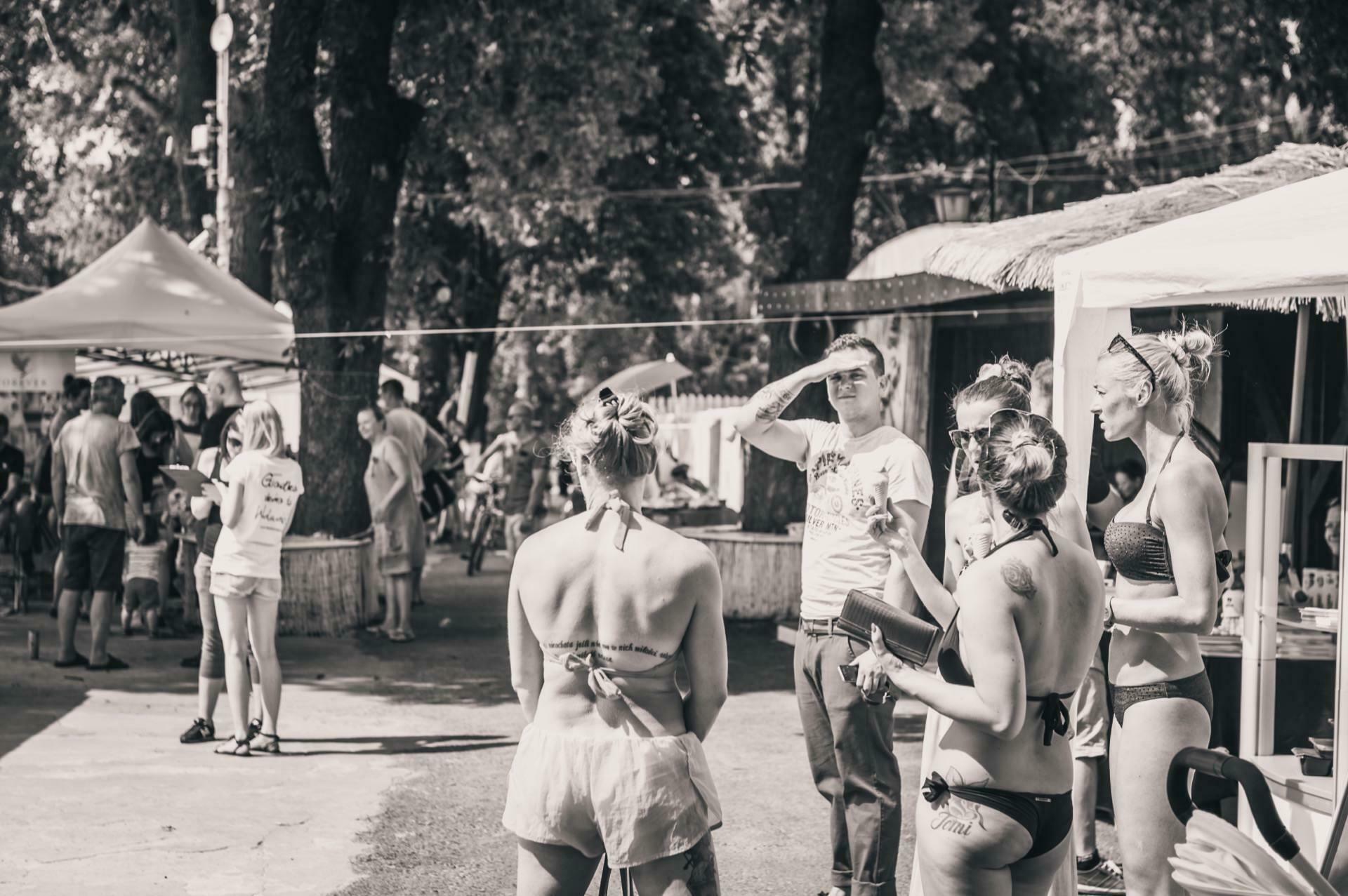 A group of people gather outdoors, dressed in casual and beach attire. Some are talking, others are standing or walking in the background. The scene includes trees, tents and a casual atmosphere, suggesting a casual or recreational event. It looks like a photo-op of a trade show.   