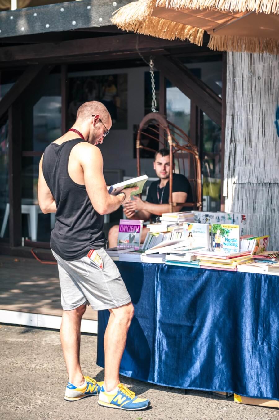 Mężczyzna w czarnym podkoszulku i szarych spodenkach przegląda książki przy księgarni na świeżym powietrzu, co przypomina ożywioną fotorelację z targów. Kolejna osoba siedzi za ladą pod parasolem krytym strzechą. Na niebieskim obrusie wyeksponowano różne książki z kolorowymi okładkami.