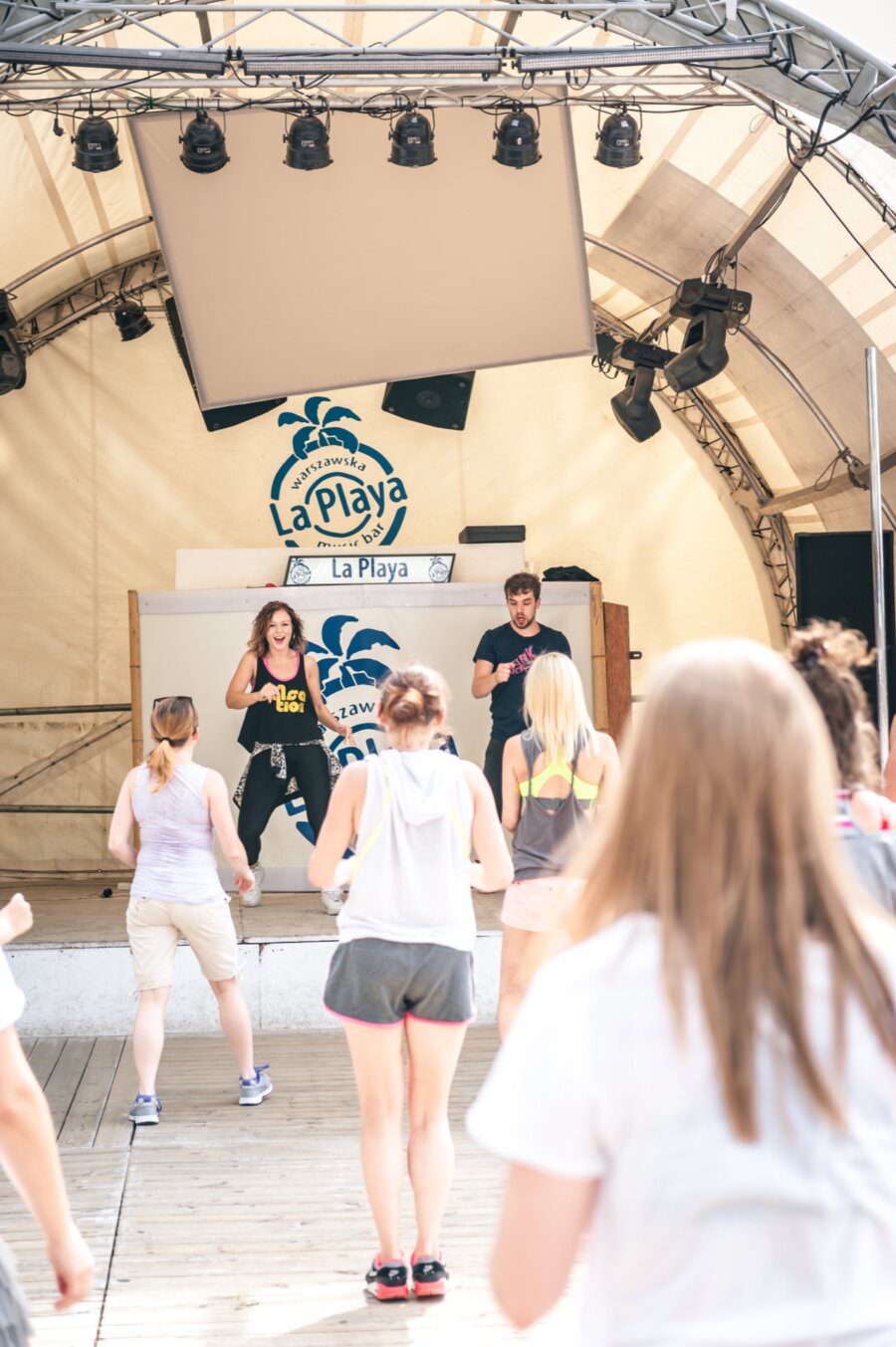Grupa osób uczestniczy w zajęciach taneczno-fitness na drewnianym tarasie pod namiotem z napisem „La Playa” w tle. Ta tętniąca życiem scena może z łatwością stać się częścią fotorelacji z targów, ponieważ dwóch instruktorów prowadzi zajęcia ze sceny, demonstrując ruchy, podczas gdy uczestnicy uważnie podążają za nimi.