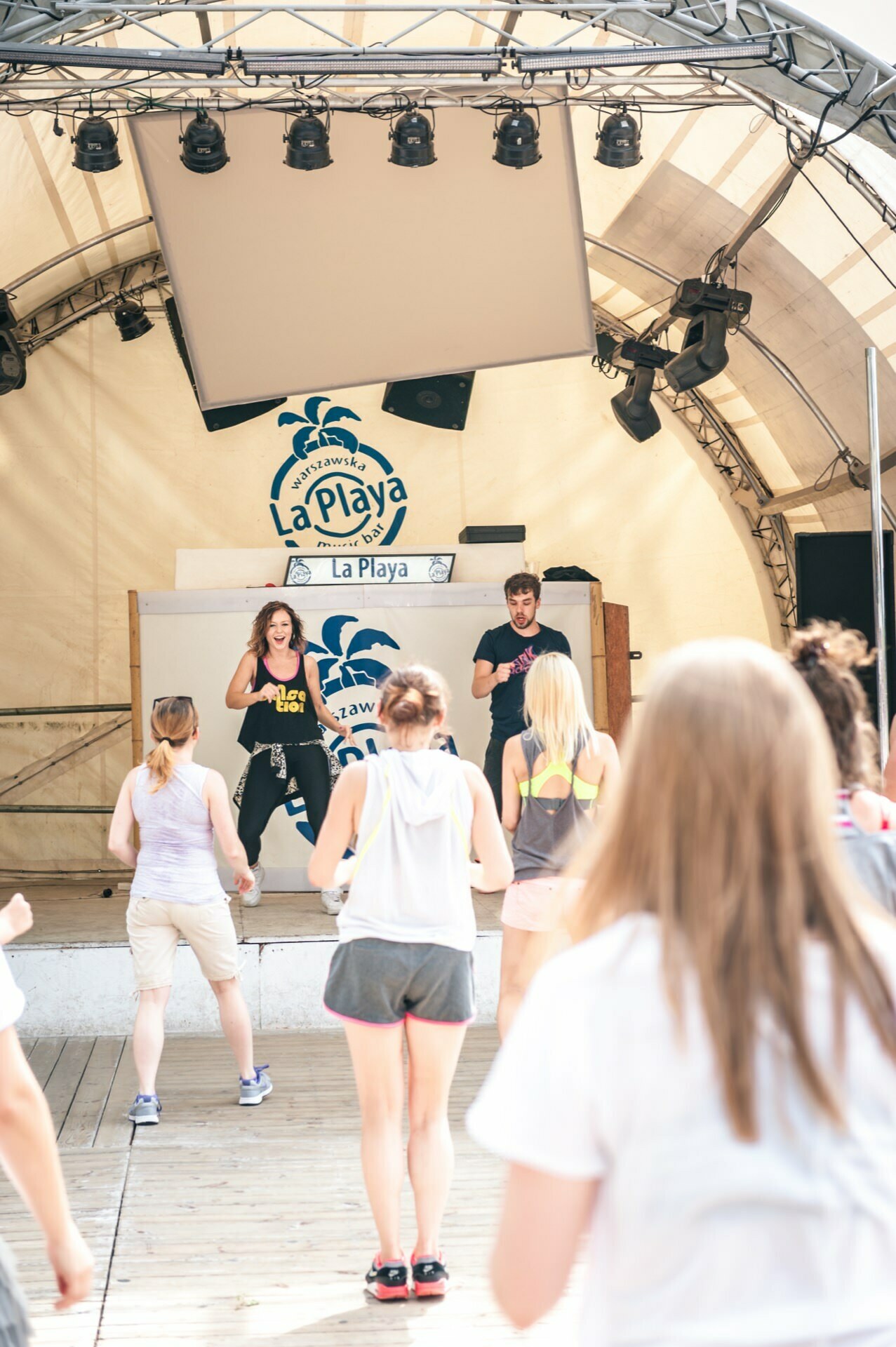 A group of people participates in a dance and fitness class on a wooden terrace under a tent with "La Playa" written in the background. This vibrant scene could easily become part of a photo essay of the fair, as two instructors lead the class from the stage, demonstrating moves while the participants carefully follow along. 