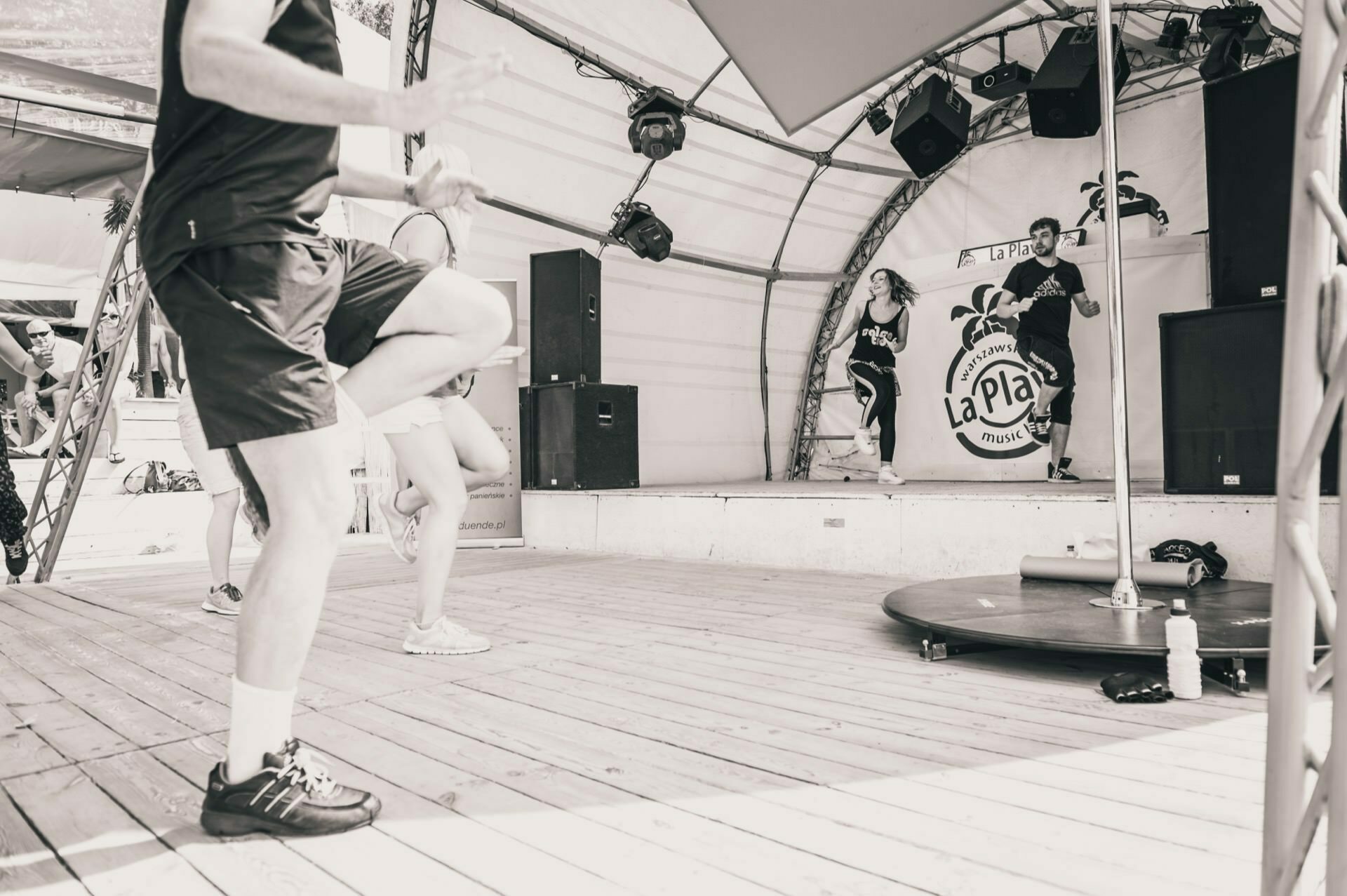 Photo report from the fair: Black and white image of a group fitness class on a wooden stage under a tent. An instructor leads a workout session while several participants perform moves. Speakers and audio equipment with a "La Playa" banner are visible in the background.  