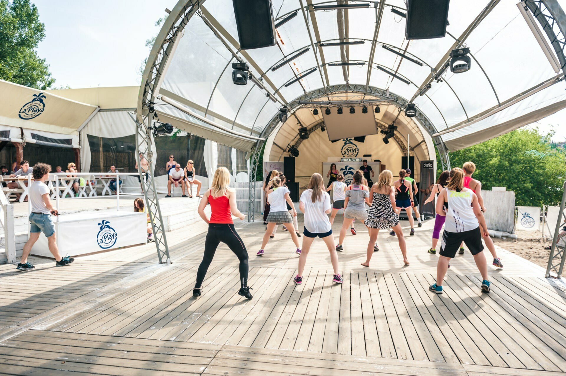 The photo report from the fair shows a group of people participating in an outdoor dance or fitness class, on a spacious wooden stage under a tent-like structure. They are dressed in sportswear and follow the movements of the instructor in front, while trees and seating areas are visible in the background. 