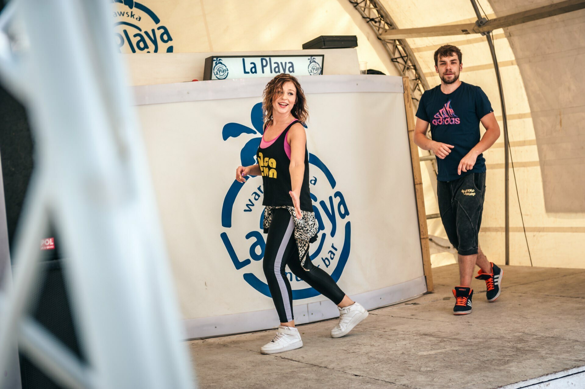 Two people are on stage at La Playa music club. The woman is smiling, wearing a black T-shirt, leggings and white tennis shoes. The man behind her is dressed in a dark T-shirt and cropped pants. Both appear to be dancing energetically. A sign on the wall in this photo of the fair reads "La Playa."    