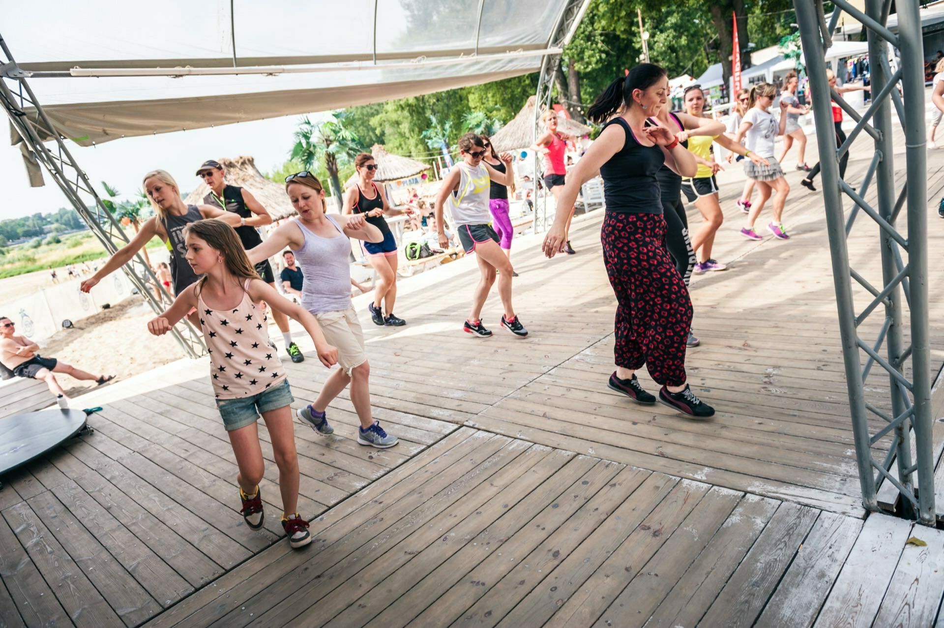 A group of people, including children and adults, take part in an outdoor dance class on a wooden stage under a canopy. They follow the instructor's directions, moving in sync. Trees and the beach can be seen in the background, capturing the vibrant atmosphere of the photo fair.  