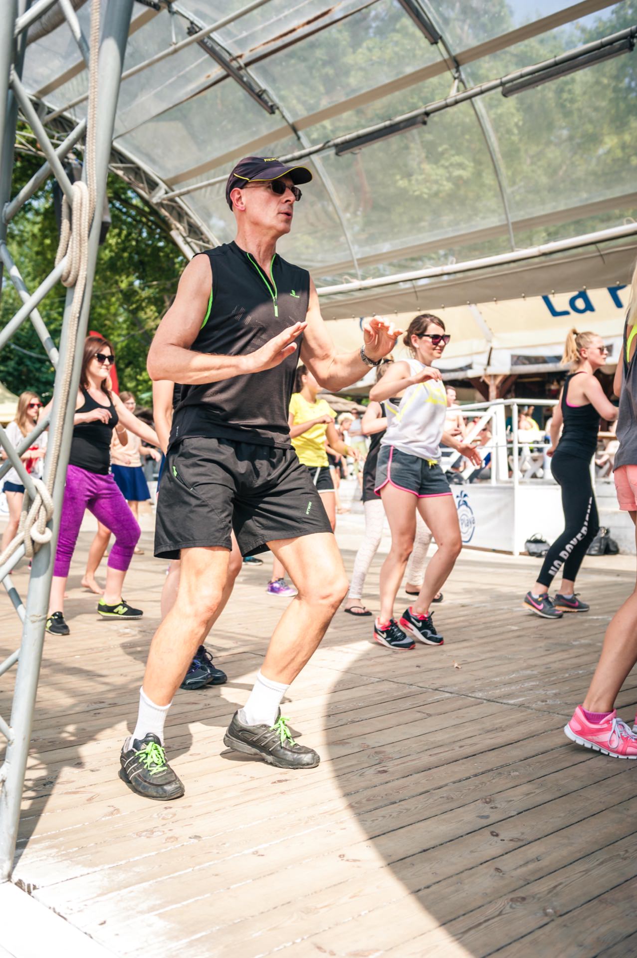 A group of people participate in a dance class or outdoor exercise on a wooden terrace under a transparent canopy. The man in the foreground is wearing a black sports outfit and sunglasses, while the others follow him according to the routine. Trees are visible in the background, reminiscent of an animated photo fair.  