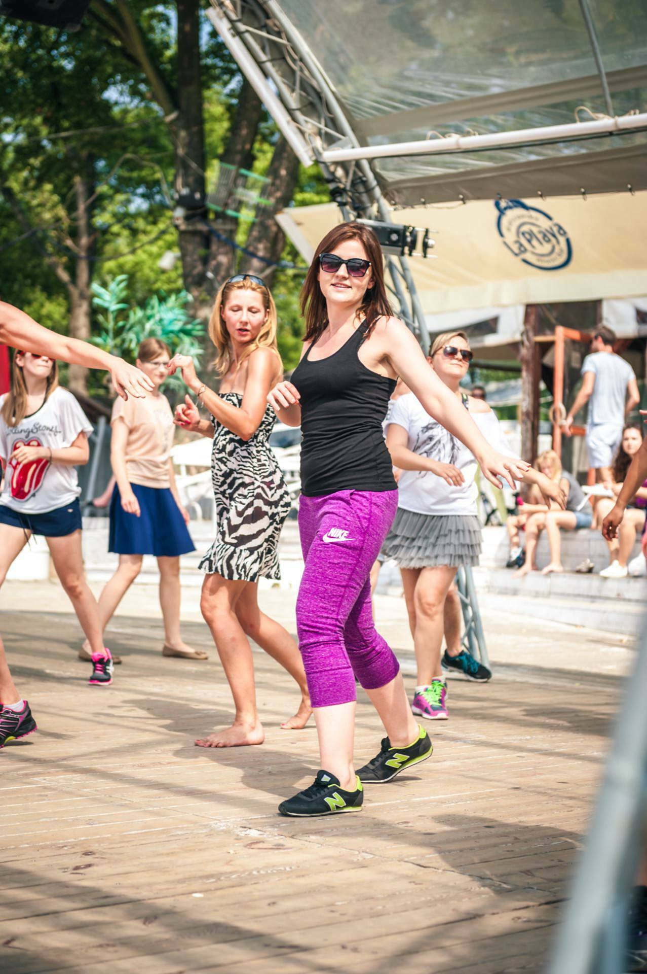 People are enjoying themselves at an outdoor dance session. The group is led by a woman wearing sunglasses, a black T-shirt and purple leggings. Others follow her movements, including women in dresses and casual outfits. The event, reminiscent of a live photo-op, takes place under a canopy with trees and a building in the background.   