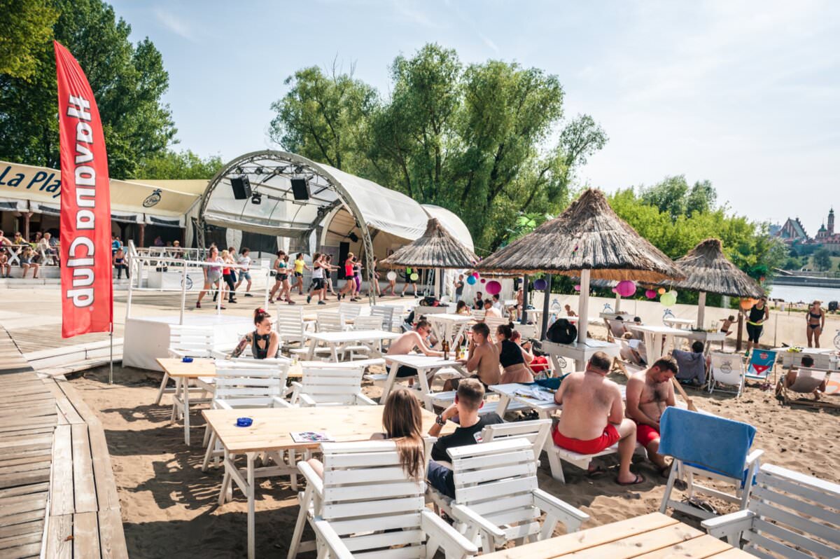 Ludzie odpoczywają na słonecznej plaży z białymi leżakami i drewnianymi stołami. W tle widać scenę z namiotem i słomianymi parasolami. Niektórzy siedzą i rozmawiają, podczas gdy inni spacerują. Po lewej stronie widoczna jest czerwona flaga z białym tekstem, jak fotorelacja z targów.