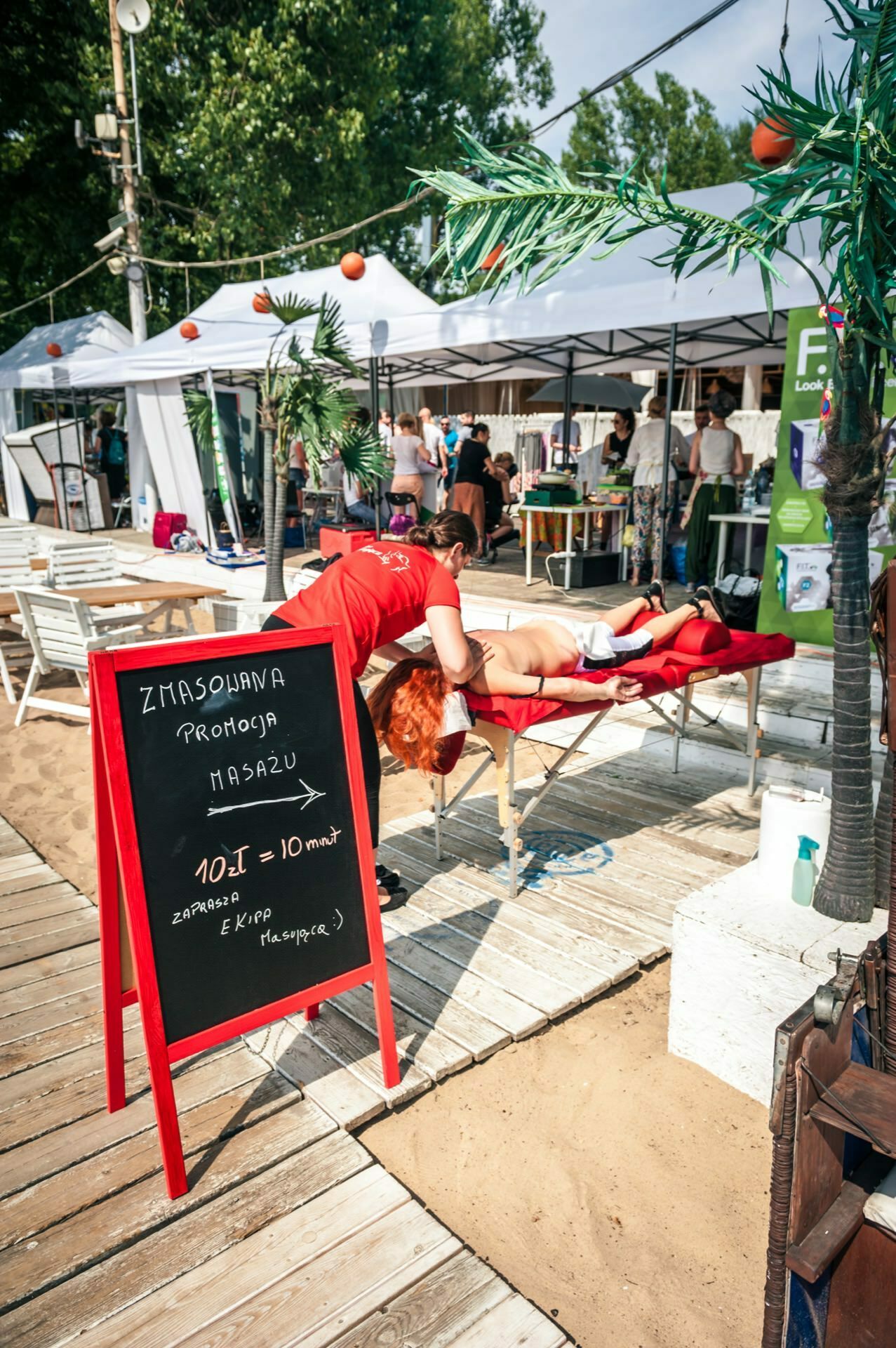 In this tranquil photo essay from the fair, a person enjoys a soothing massage on a red table by the beach. A black board sign with white and orange text stands out nearby. People stroll under the white tents in the background, accompanied by lovely palm tree decorations.  