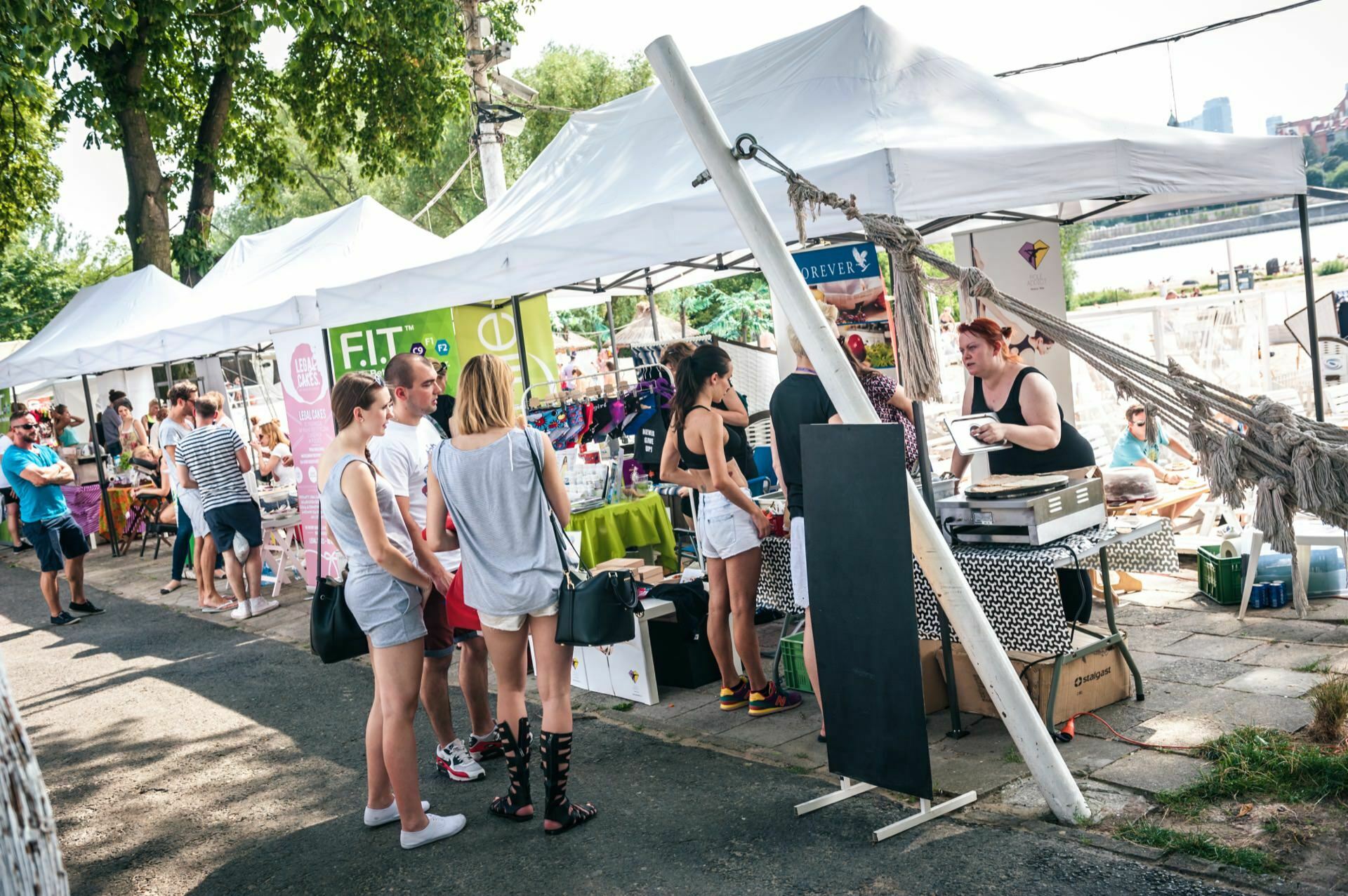 A group of people dressed in ordinary summer clothes are browsing market stalls with white tents. Vendors display a variety of goods on tables, while bags pile up underneath. Trees and a river can be seen in the background, suggesting a bustling, open-air market by the water - Photo Report from the fair.  