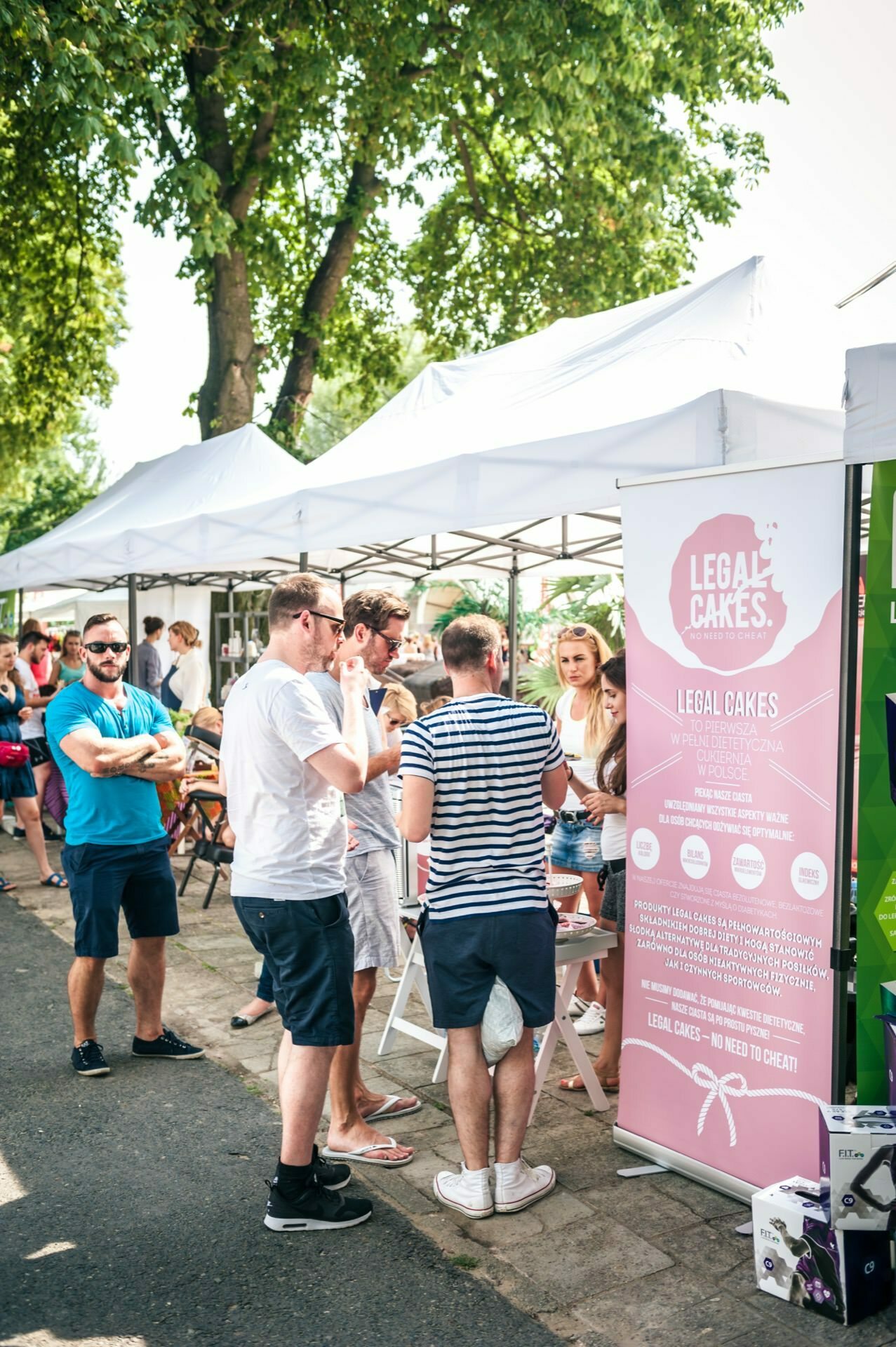 A group of people stand in front of a stall with a pink banner reading "LEGAL CAKES." The stall with a white canopy is set up along a tree-lined street. On a sunny day, people are chatting and enjoying the atmosphere of the open-air market - the perfect place for a photo report of the fair.  