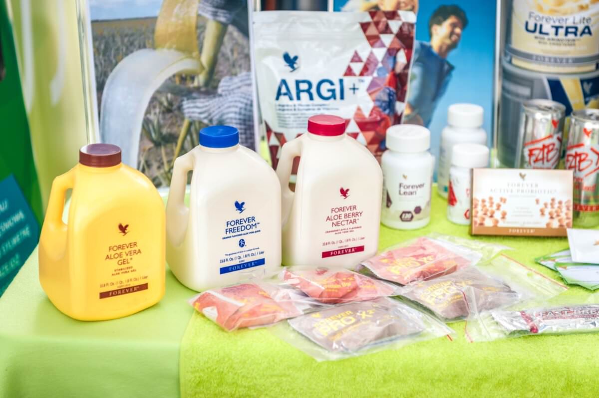 Ekspozycja różnorodnych produktów zdrowotnych i wellness marki Forever Living. Fotorelacja z targów to duże butelki żelu Aloe Vera, nektaru z aloesu, Forever Freedom, saszetki ARGI+, butelki po suplementach diety i batony energetyczne ułożone na limonkowym obrusie.