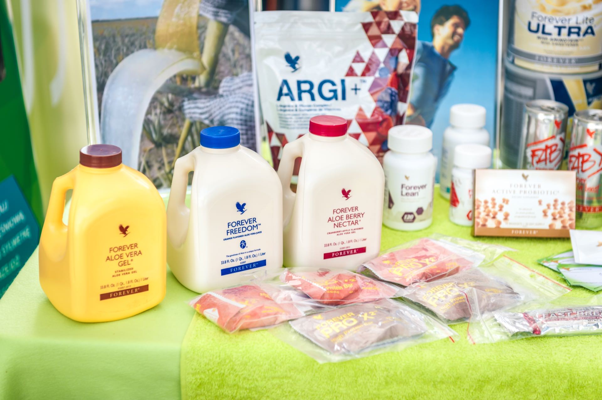 A display of a variety of health and wellness products by Forever Living. The photo exhibit features large bottles of Aloe Vera gel, aloe vera nectar, Forever Freedom, ARGI+ sachets, dietary supplement bottles and energy bars arranged on a lime green tablecloth. 