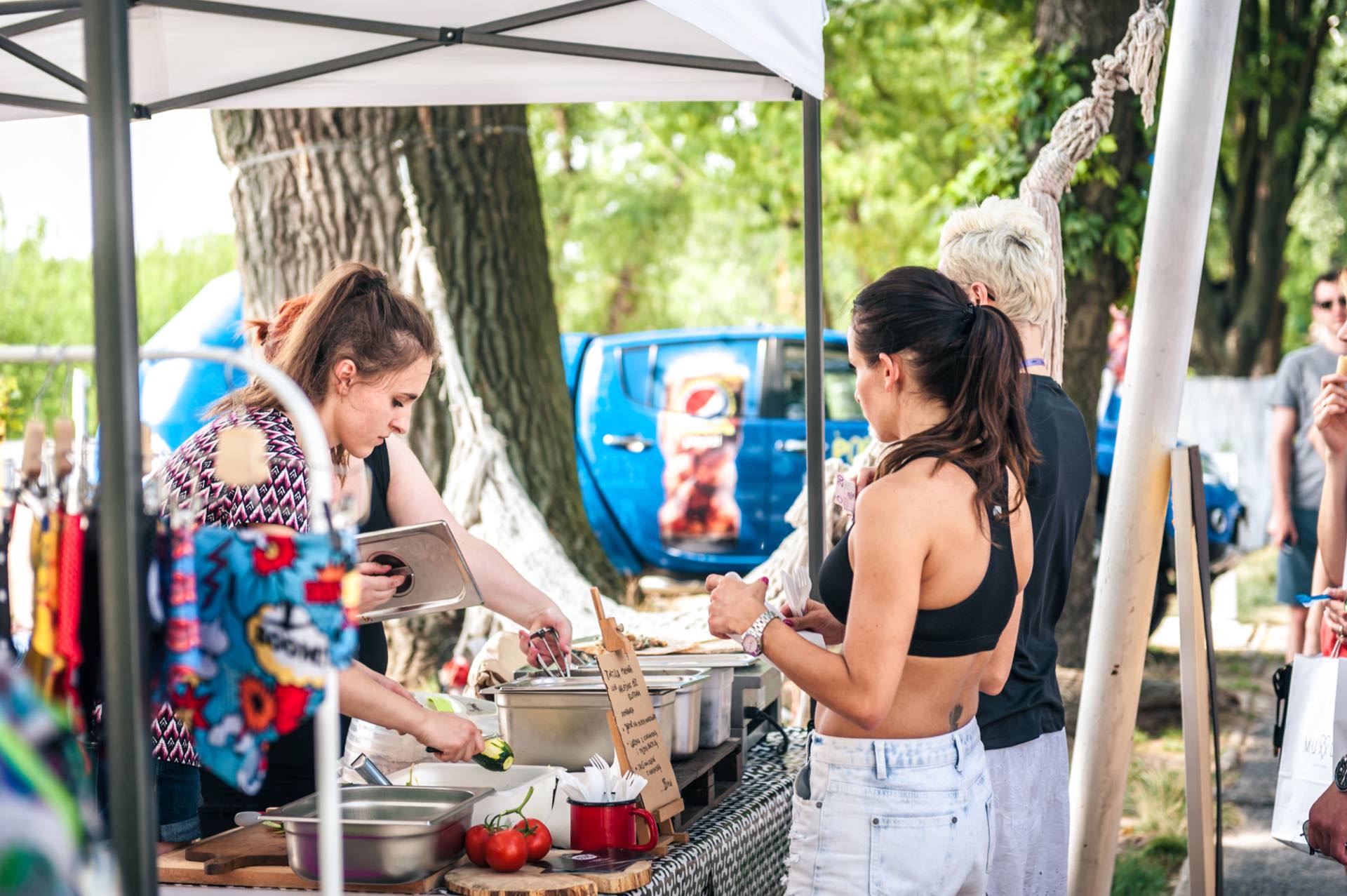 A woman in a patterned short-sleeved shirt prepares food at a stall under an outdoor canopy, while fresh produce lies on a table. The photo of the market shows two customers, one in a black T-shirt and the other in white jeans, waiting on the other side. In the background are trees and a vehicle.  