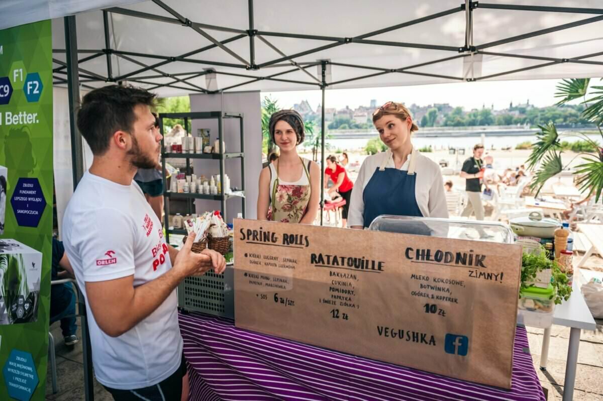 Przy stoisku z napisem „Vegushka” stoi mężczyzna z lodami i rozmawia pod baldachimem z trzema obsługującymi. W menu stoiska znajdziemy sajgonki, ratatouille oraz zimne napoje. Obsługa uśmiecha się, obserwując wydarzenie na świeżym powietrzu w pobliżu plaży – idealne dla naszej fotorelacji z targów.