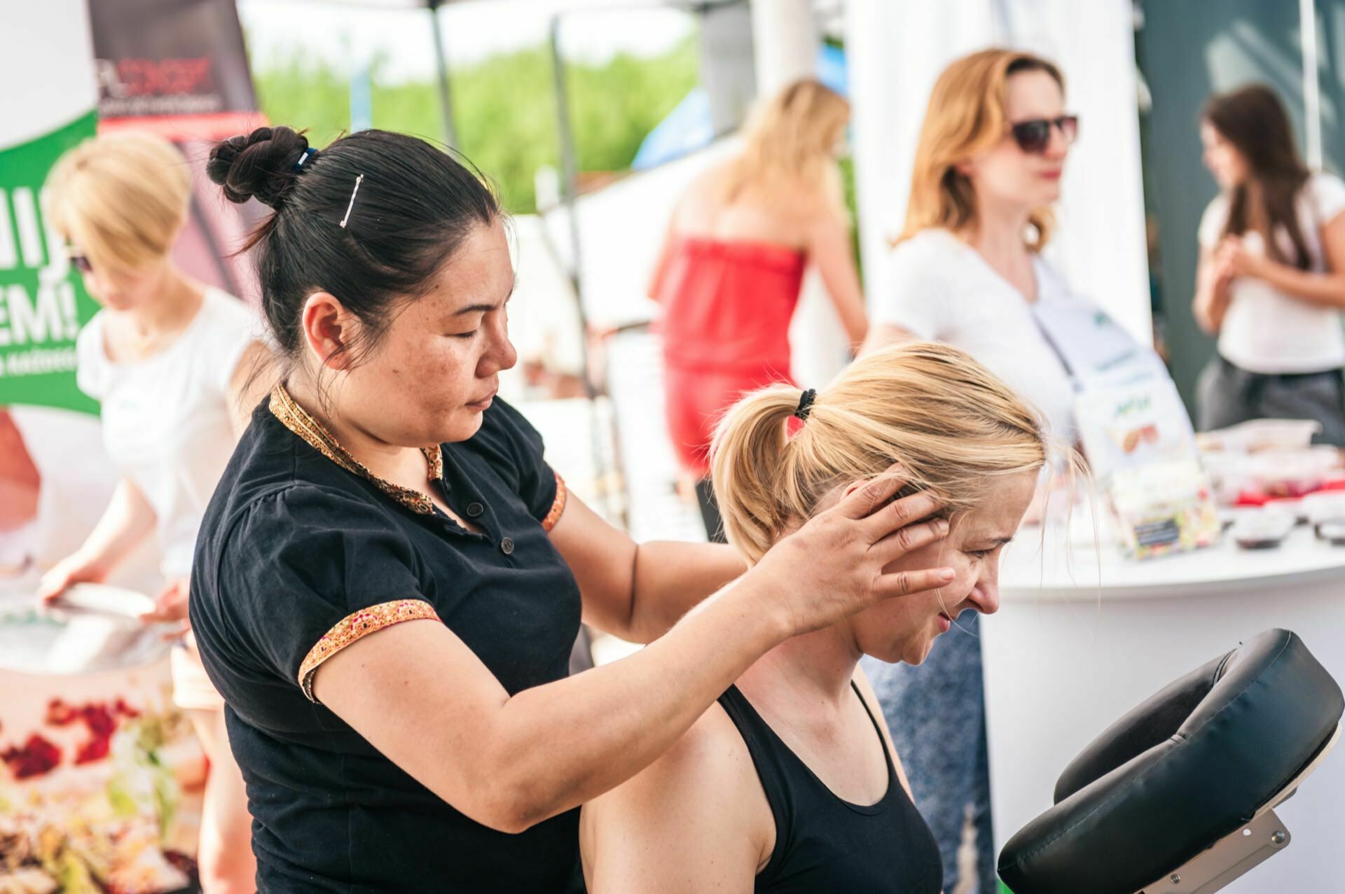 A woman undergoes a neck and head massage in a sunny outdoor setting as part of a photo fair, surrounded by other people busy with various activities. The masseur uses both hands to apply pressure, while the client sits and looks relaxed. 