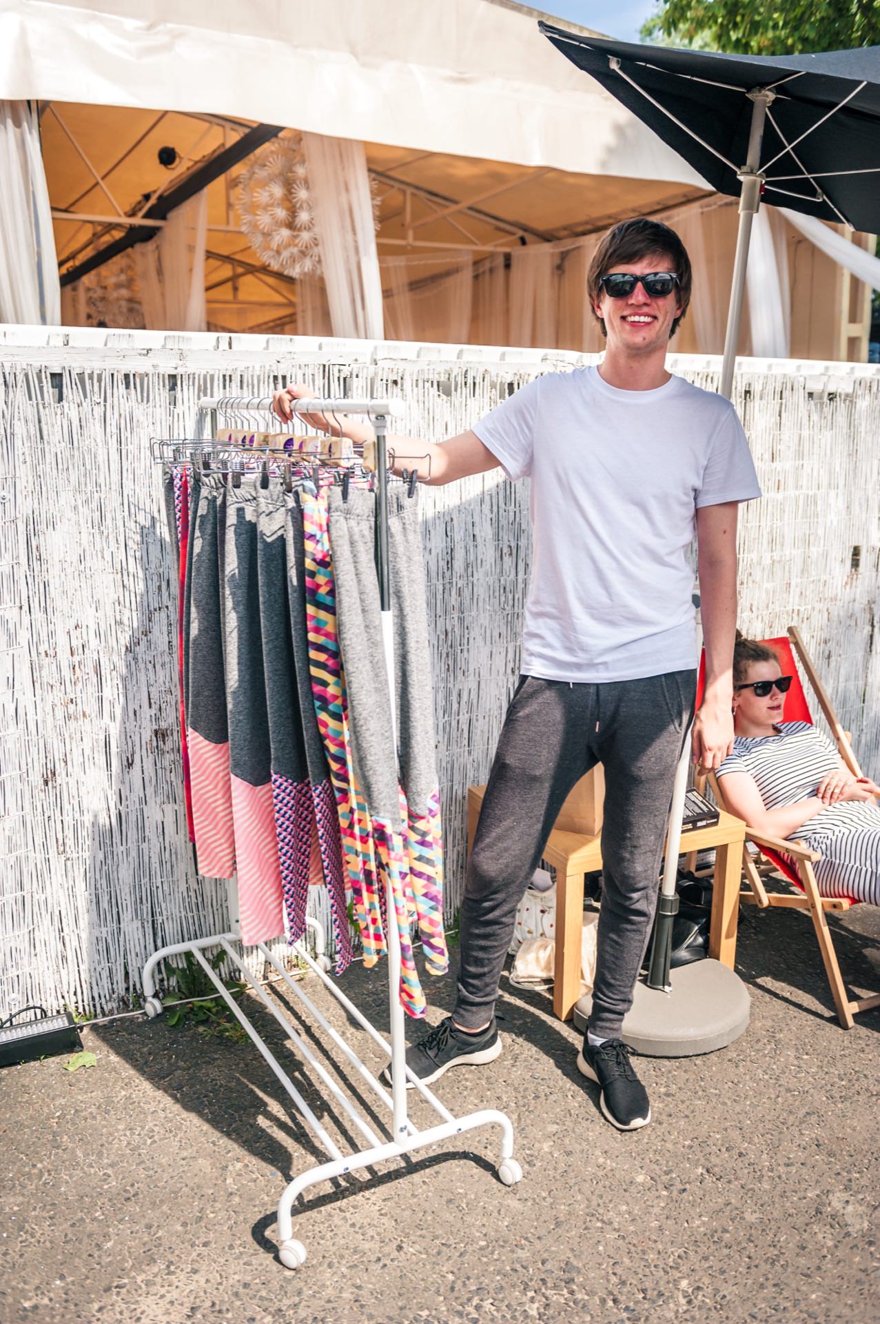 A person in a white t-shirt and dark pants is standing and smiling next to a clothing rack on which a variety of colorful pants are posted, outside the outdoor seating area. Another person, captured in our photo report from the fair, is lying on a deck chair in the background, wearing a striped dress and sunglasses. 