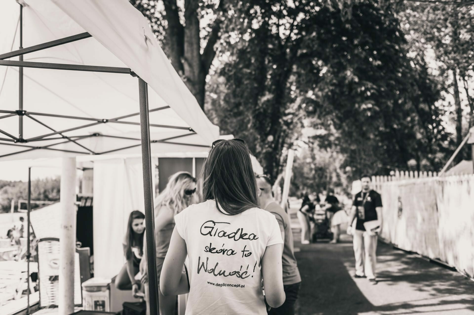 Black and white photo of a person with long hair walking away under a canopy. They are wearing a T-shirt with text on the back. The busy outdoor atmosphere captured in this photo essay from the fair is a few people and a fence with trees in the background.  