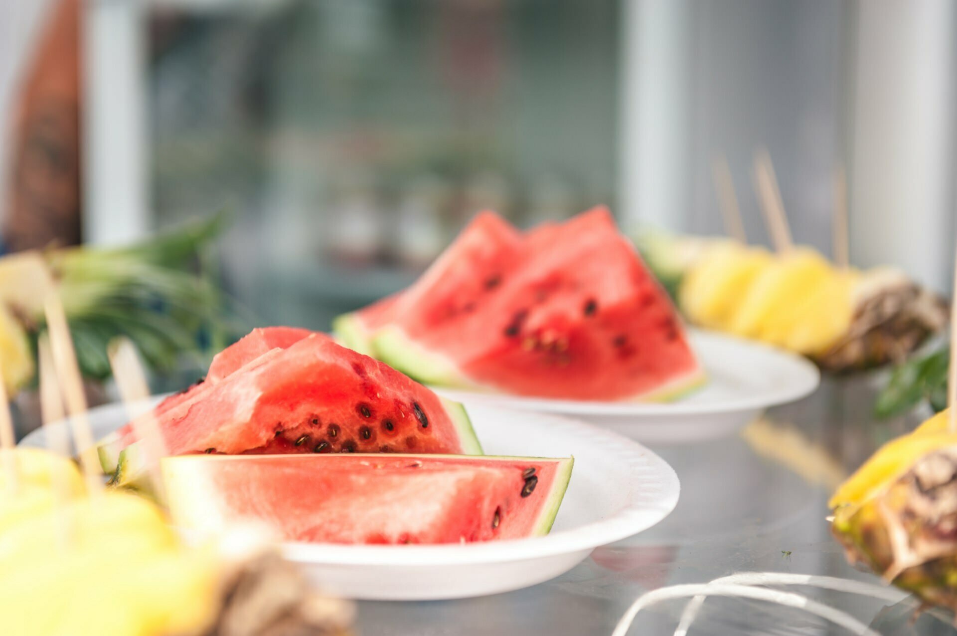 Slices of fresh watermelon and chunks of pineapple on skewers are arranged on white plates in a bright setting, reminiscent of a photo fair. The background is blurred to emphasize the vibrant colors and juicy texture of the fruit in the foreground. 