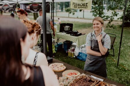 A woman dressed in a gray apron stands smiling behind a stall at an outdoor market in Warsaw. The stall features a wide variety of food products, including pies and large cakes. Above her was a sign reading "Pierogi and other delicacies." In the back, several people can be seen waiting in line, while an event photographer captures the moment.   