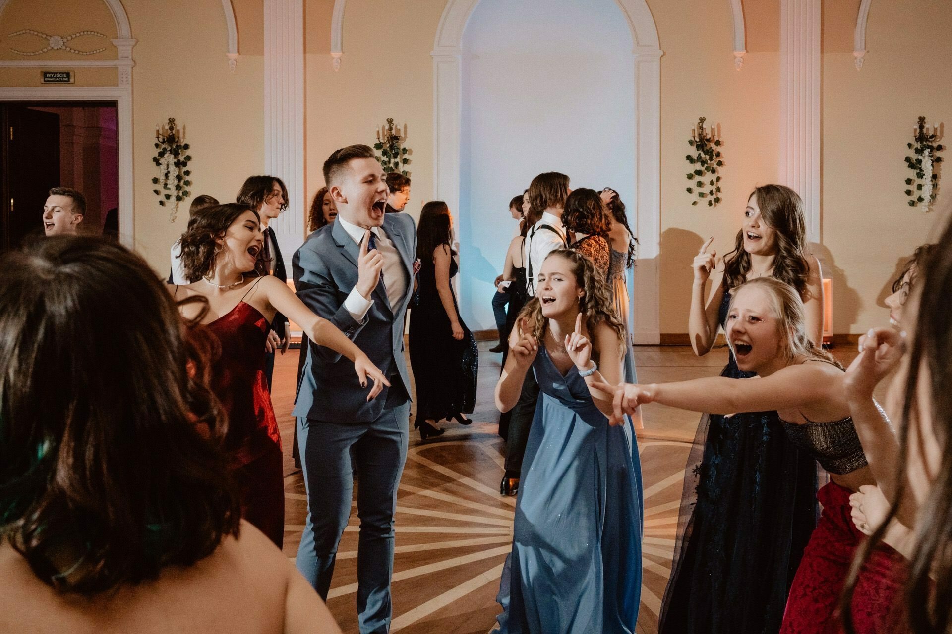 A group of formally dressed young people joyfully dance and sing at a hall party reminiscent of a prom reportage. In the background are elegant arches and decorations in the form of greenery. It looks like everyone is having a great time, as can be seen by the animated faces and lively gestures.  