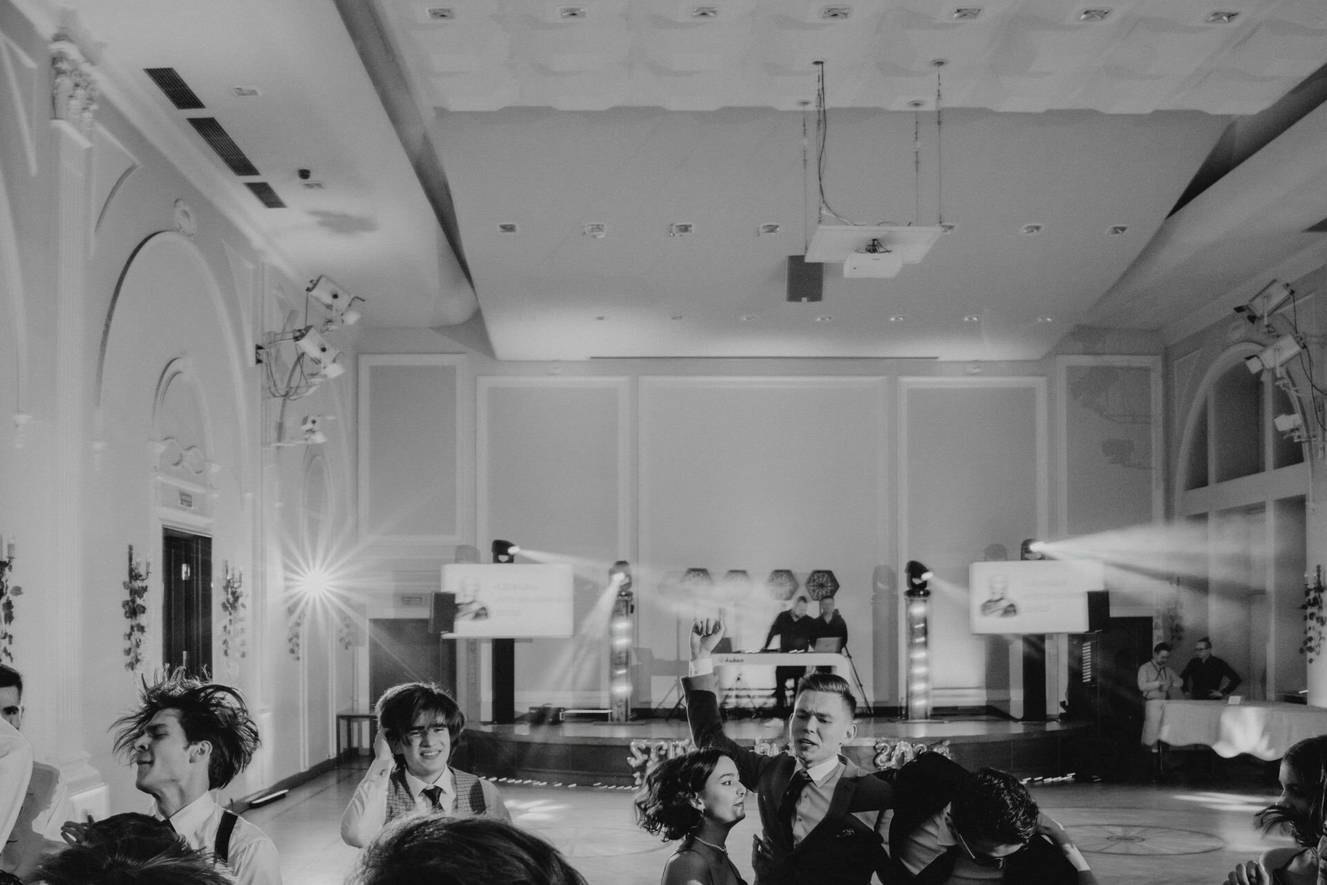 A black-and-white photo of a dimly lit banquet hall shows a DJ playing music in what looks like a report from a prom. Attendees are dancing on the floor, some laughing and talking. The room has high ceilings, arched doorways and ornate light fixtures. Bright spotlights illuminate the DJ area.   