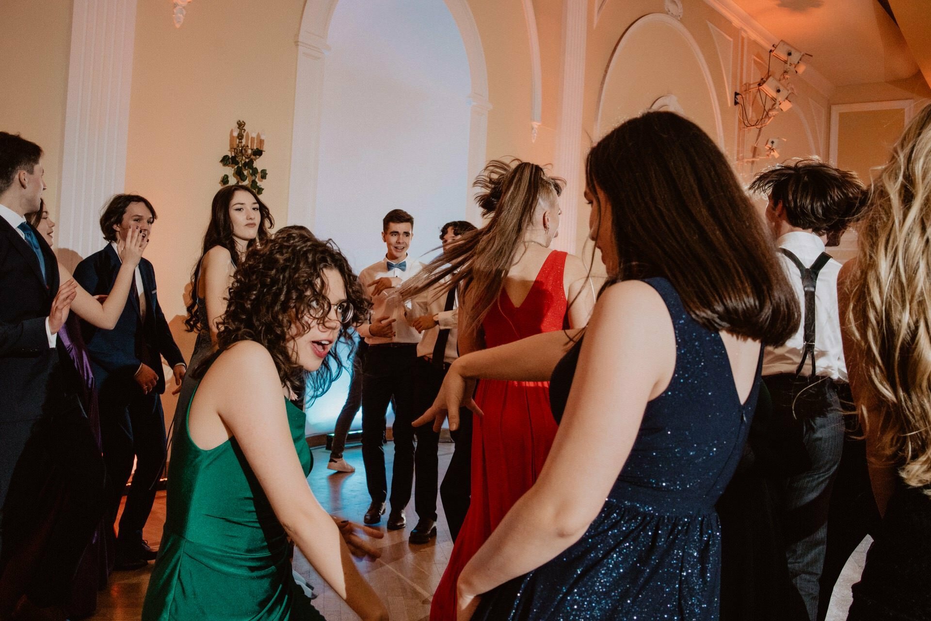 A group of young adults in formal attire dance in a grand ballroom with tall white columns and arched doors. The vibrant scene, reminiscent of a prom reportage, includes people in dresses and suits enjoying the music and atmosphere, while warm lighting illuminates the space. 