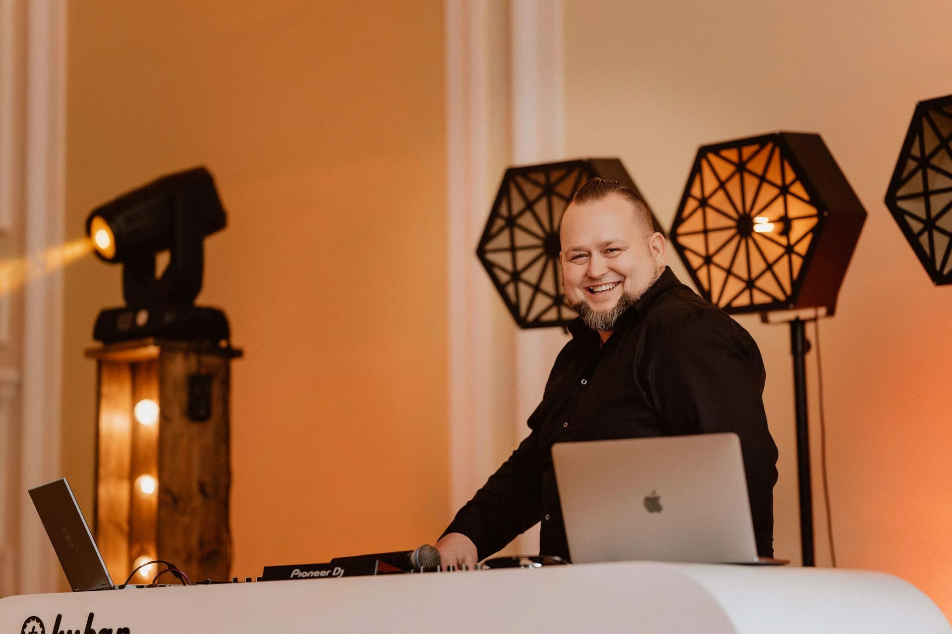 A DJ in a black shirt stands behind a white DJ booth with equipment and a laptop and smiles. The scene could resemble a report from a prom, with modern geometric lamps and stage lighting adding to the vibrant atmosphere. 