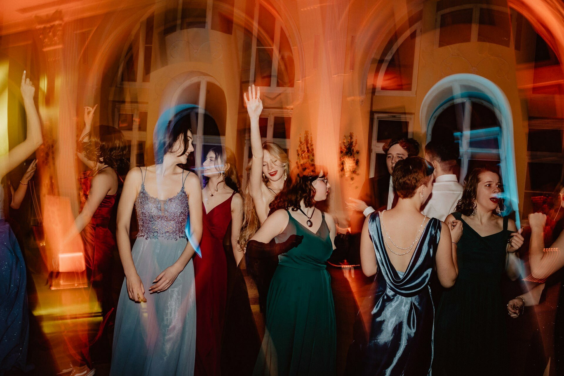 A group of people in formal attire dance vigorously in a dimly lit room with large arched windows. The colorful lights create a fuzzy, dynamic effect around the dancers, capturing the essence of a prom reportage. Some have their hands raised and everyone seems to be enjoying the vibrant atmosphere.  