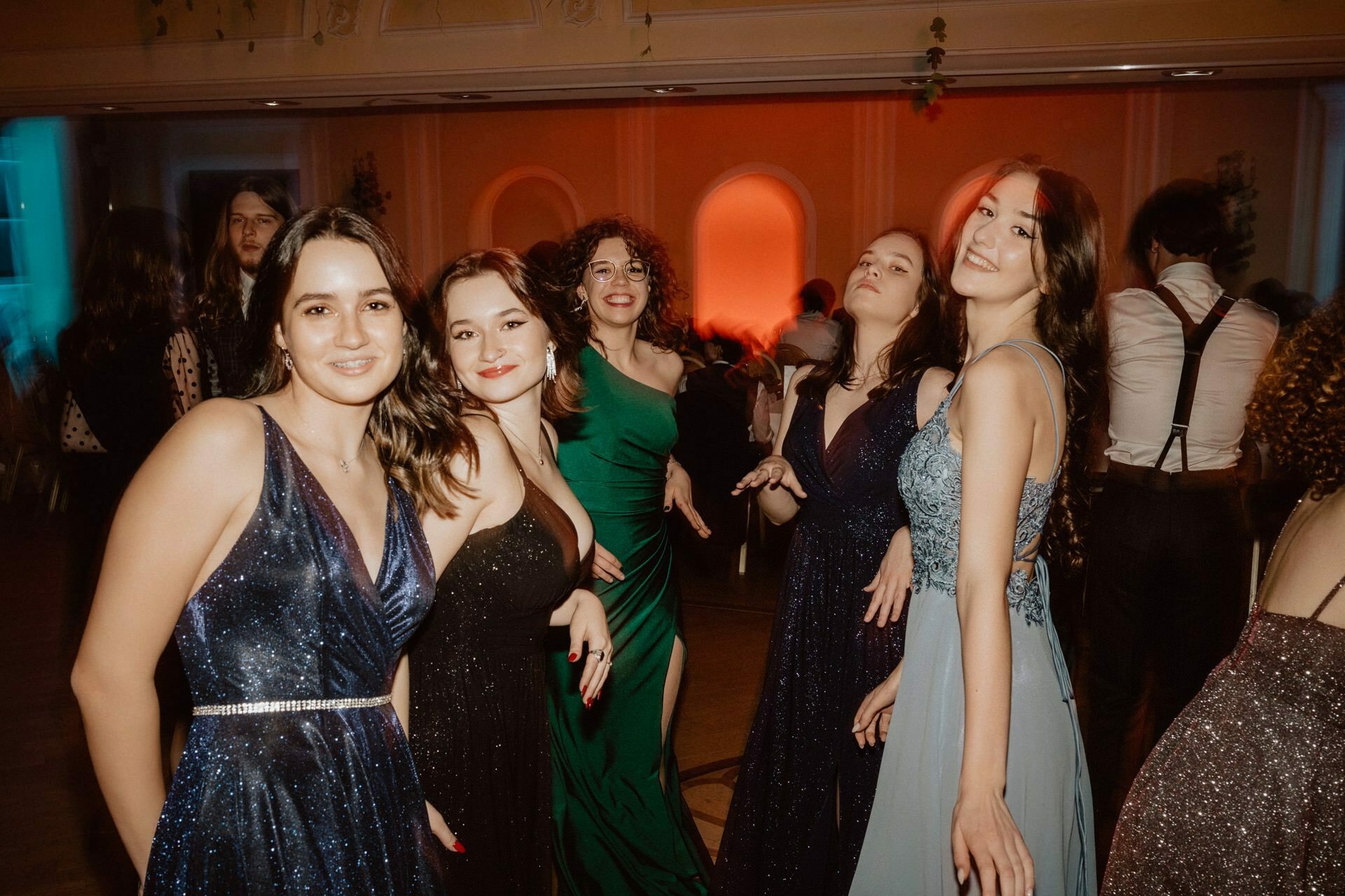 A group of five young women in formal gowns smile and pose together during a lively hall event, reminiscent of a bustling prom reportage. The background is characterized by warm lighting and mingling people. Two women are wearing blue dresses, one in black, one in green and one in a light blue dress.  