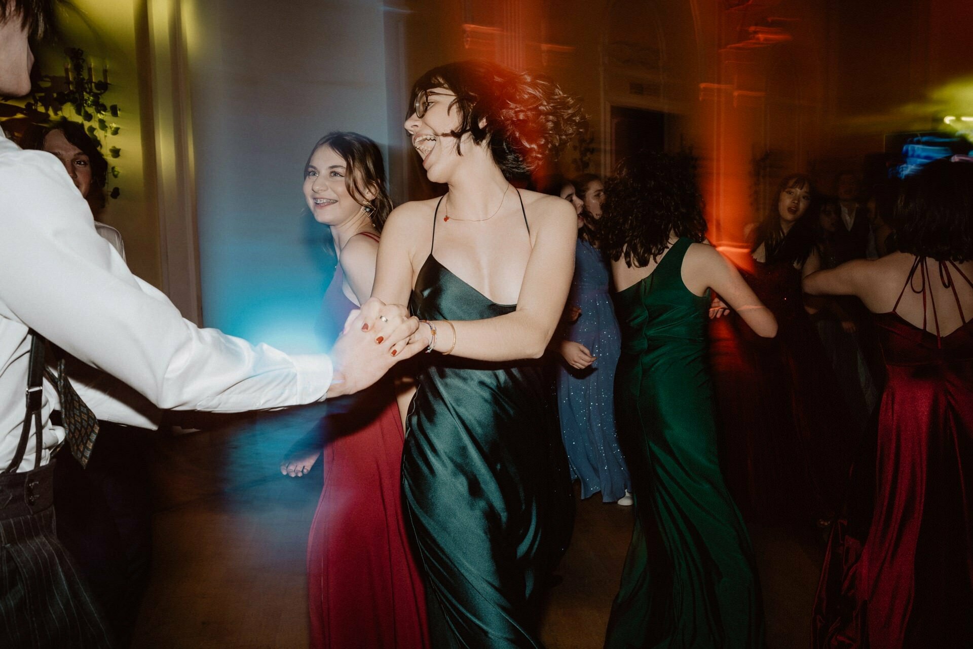 A group of people in formal attire are dancing in a dimly lit room with colored lights, reminiscent of a prom reportage. In the foreground stands a woman in a green dress, holding hands and dancing vigorously with someone in a white shirt. Others in the background are also dancing and smiling.  