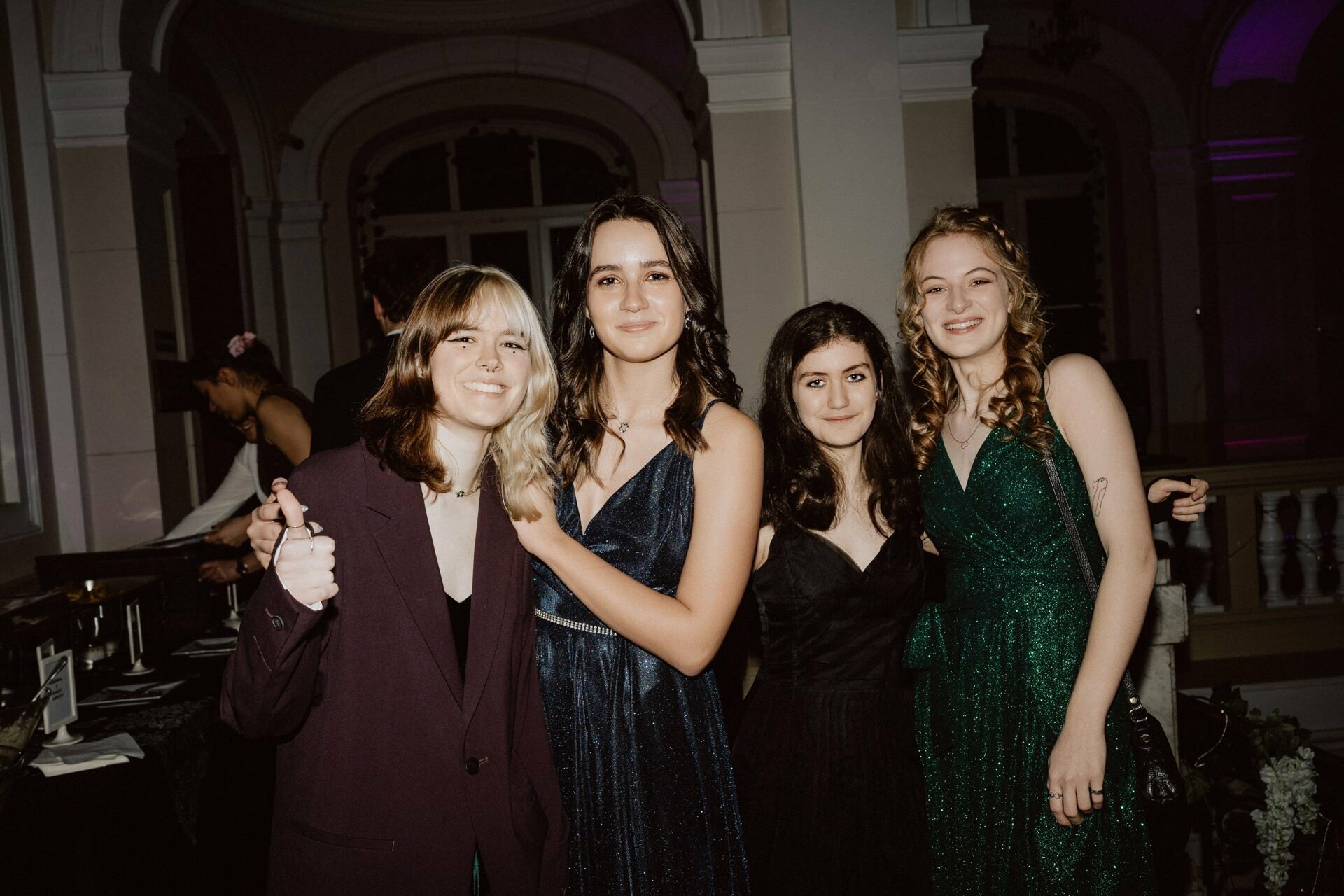 Four young women dressed in formal attire smile as they pose together at the event. One woman on the left has a two-tone hairstyle and a maroon suit, showing thumbs up. The other three, in dresses in shades of blue, black and green, stand in the decorated hall - a perfect snapshot of a "prom reportage."  