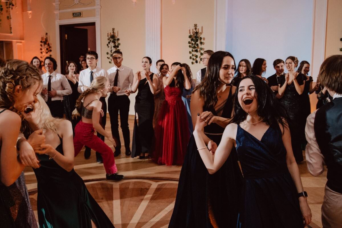 Grupa osób ubranych w formalne stroje tańczy i spotyka się towarzysko w dużej, elegancko urządzonej sali. W tętniącym życiem reportażu ze studniówki kilka osób na pierwszym planie energicznie tańczy, inne w tle są pochłonięte rozmową i obserwowaniem tancerzy.