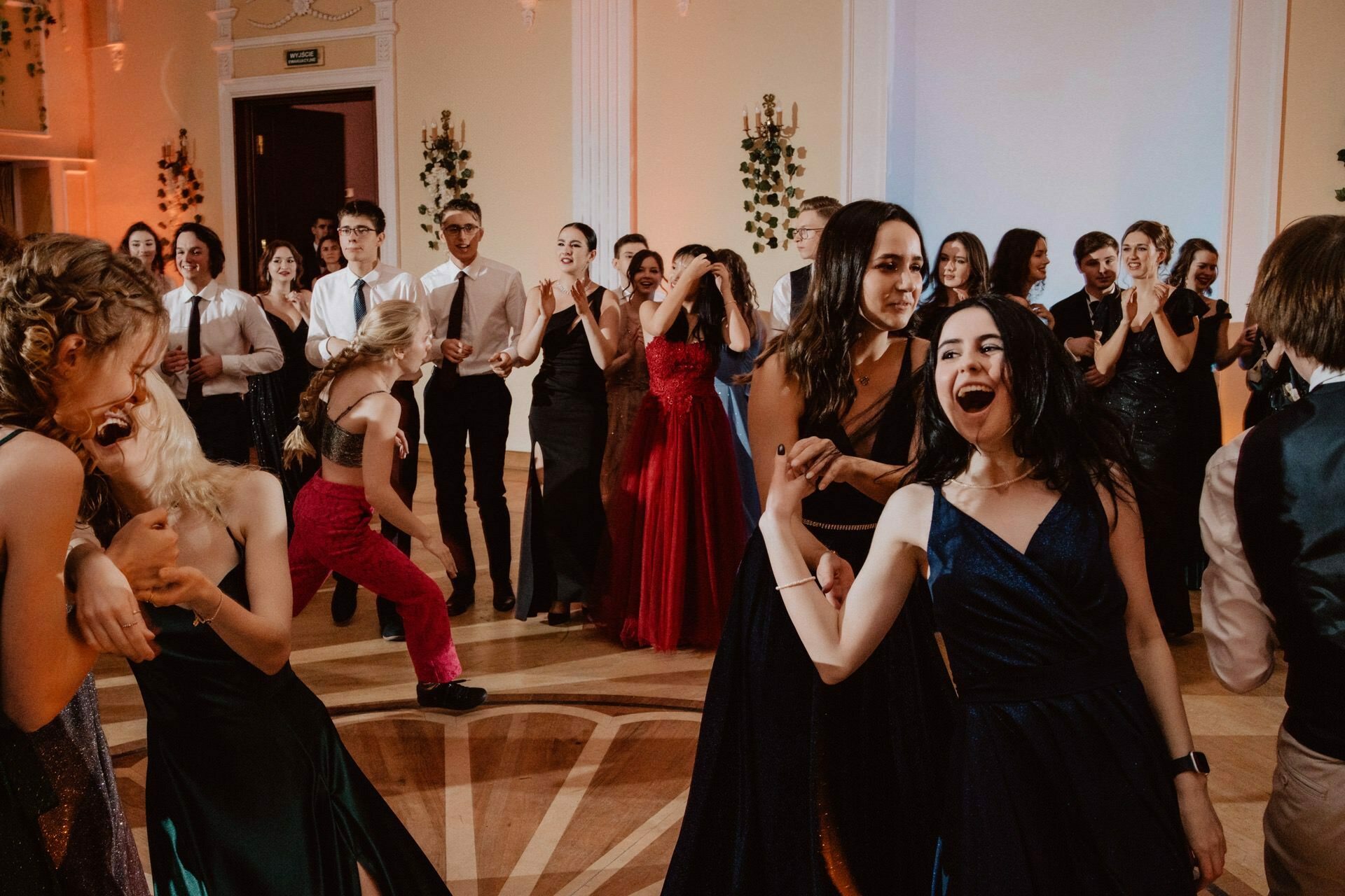 A group of people dressed in formal attire dance and socialize in a large, elegantly decorated hall. In a lively reportage of the prom, a few people in the foreground are vigorously dancing, while others in the background are engrossed in conversation and watching the dancers. 