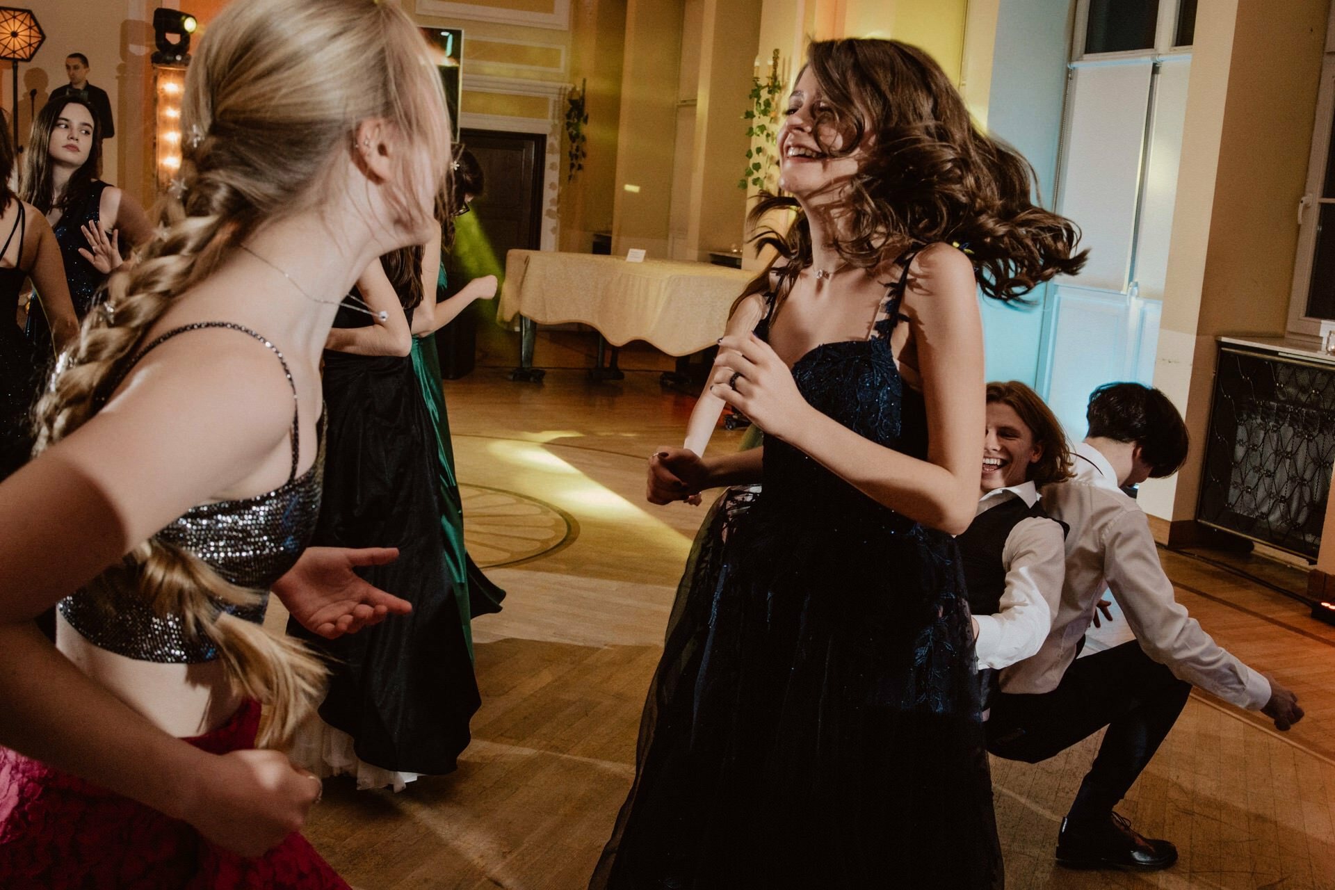 A group of well-dressed young people dance energetically in a well-lit room with wooden floors. In the foreground two girls, one with braids and the other with loose hair, are smiling and having fun. One has the impression of a vibrant reportage of a prom, while others are dancing and laughing in the background.  