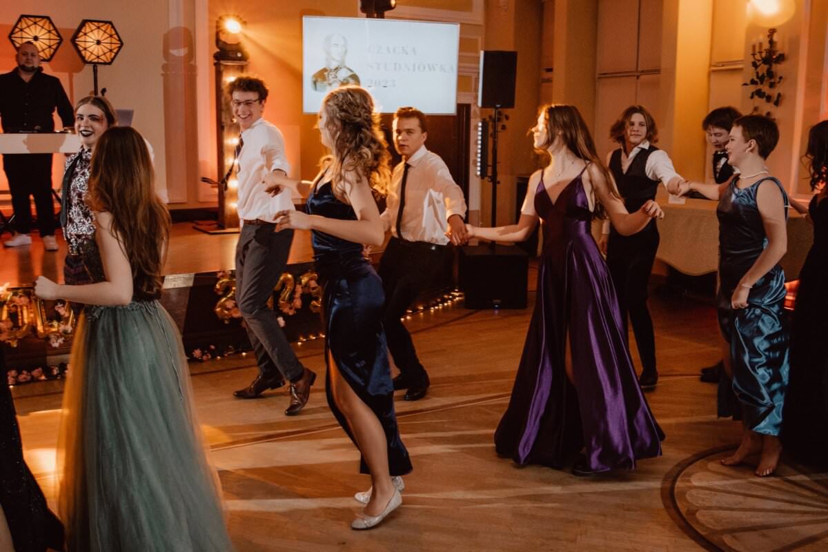 Grupa osób w strojach formalnych tańczy w dobrze oświetlonej sali balowej. Kobiety noszą eleganckie suknie, a mężczyźni – koszule i spodnie. Sala ma ciepłe oświetlenie, a stanowisko DJ-a ze świecącymi dekoracjami to idealne miejsce na Reportaż ze studniówki.