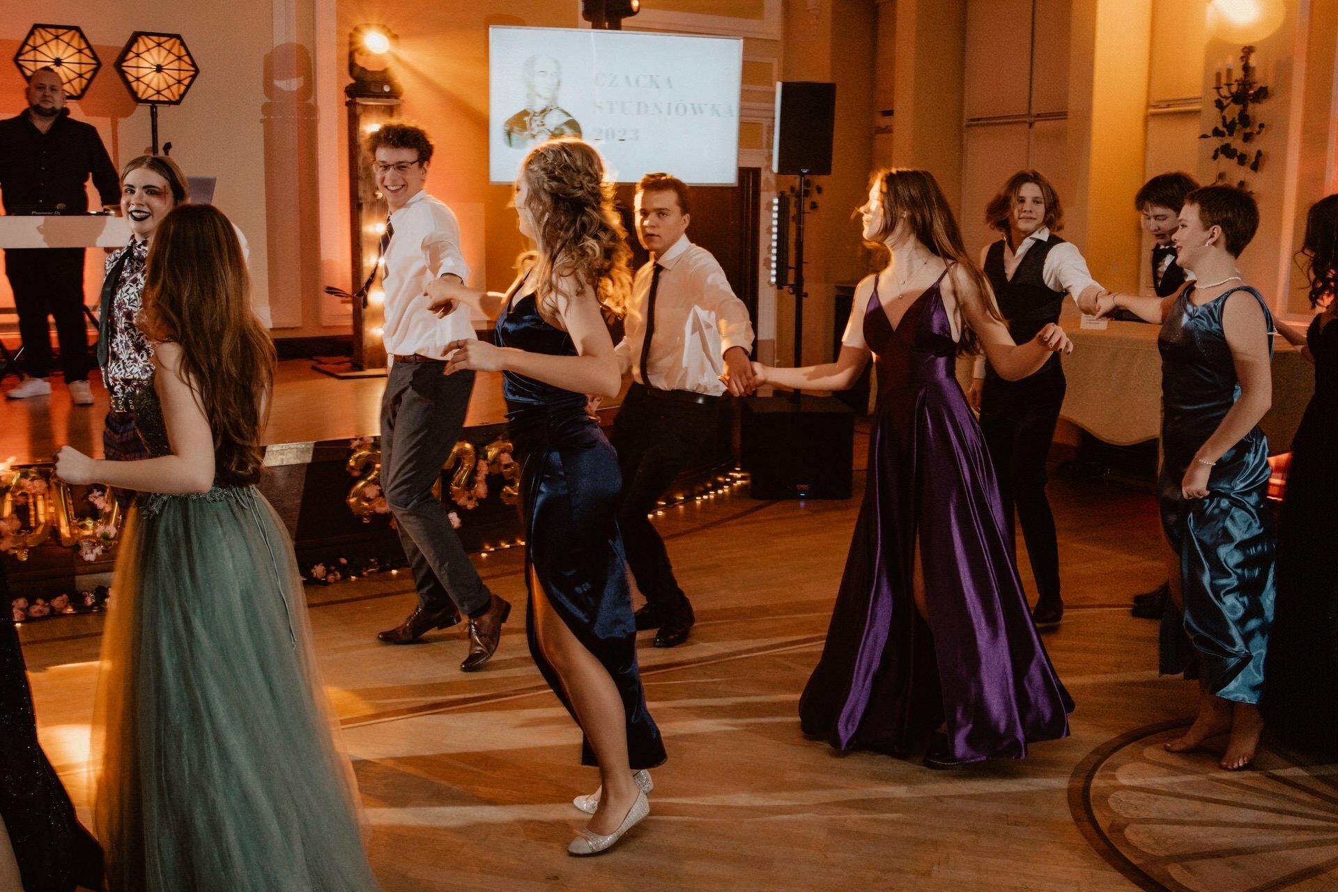 A group of people in formal attire dance in a well-lit ballroom. Women wear elegant gowns, while men wear shirts and pants. The hall has warm lighting, and a DJ booth with glowing decorations is the perfect place for a Prom Report.  