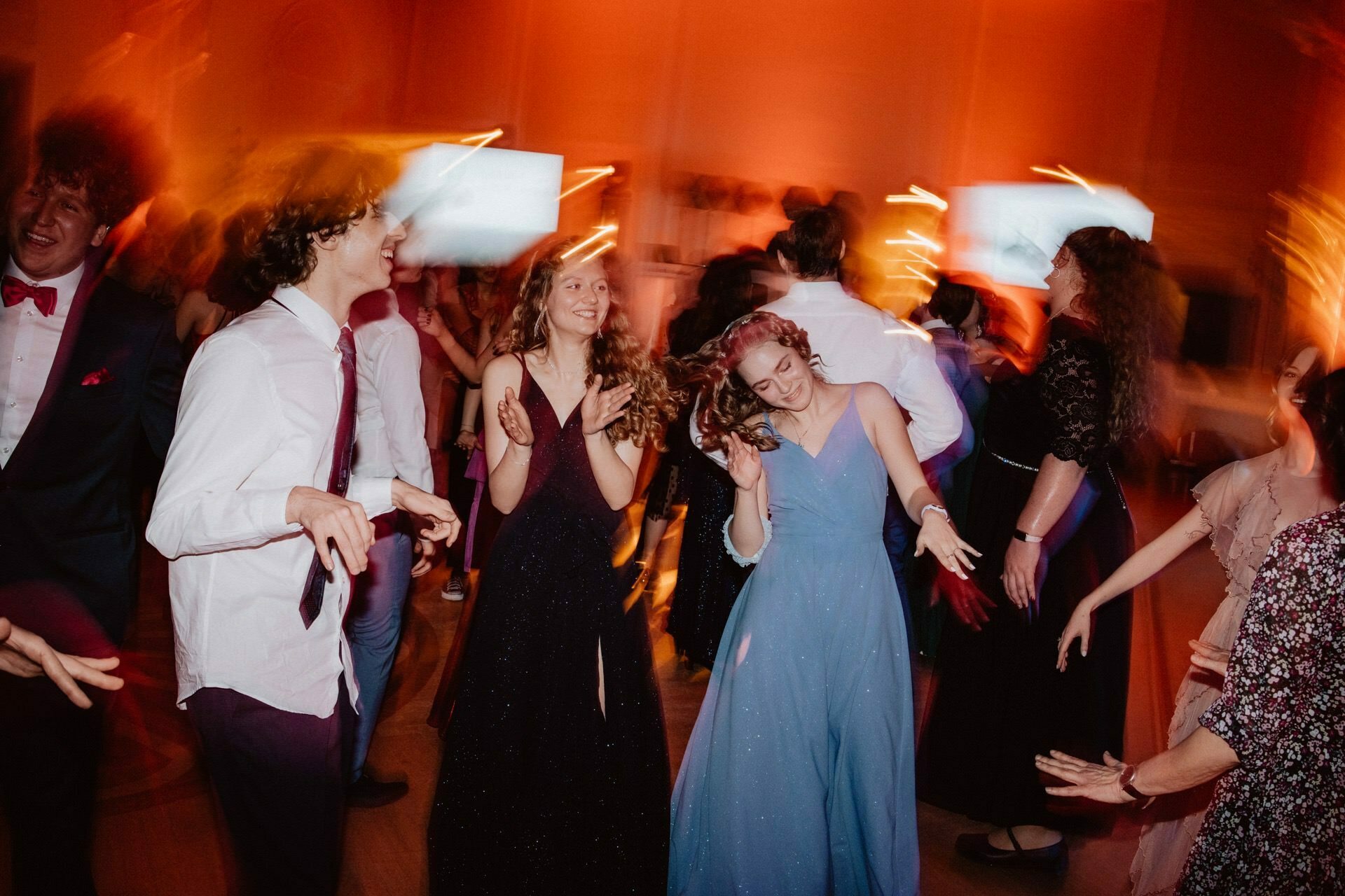 The prom reportage captured a lively scene of young people dancing at a social event. They are dressed in formal attire, women in gowns, men in shirts and ties. The lighting is warm and colorful, creating a lively atmosphere. The motion blur adds to the impression of movement and excitement.   