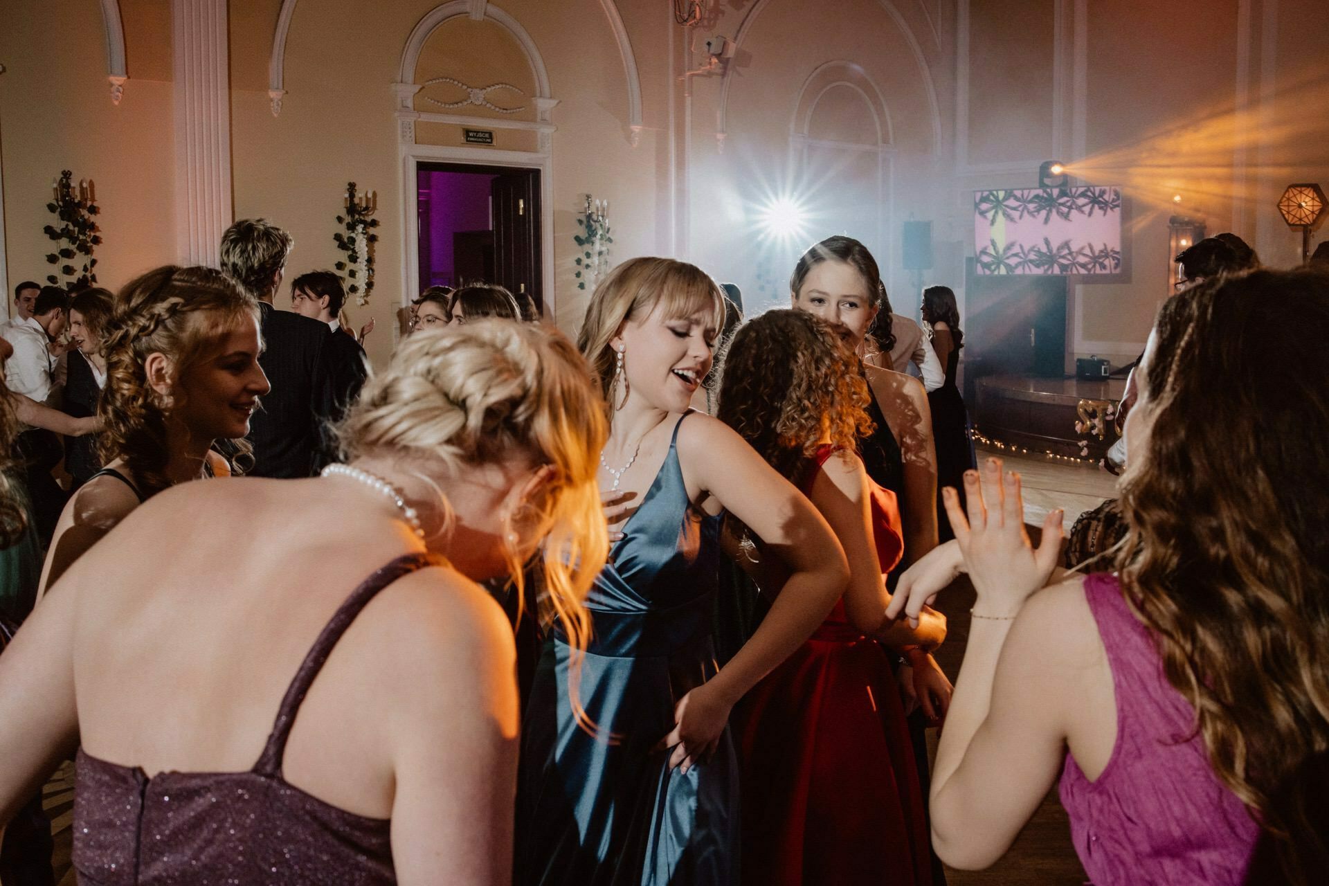 A group of people dressed in formal attire dance in a decorated hall. Women dressed in multi-colored gowns turn up the mood. Warm lighting and colorful effects add to the festive atmosphere of the prom reportage.  