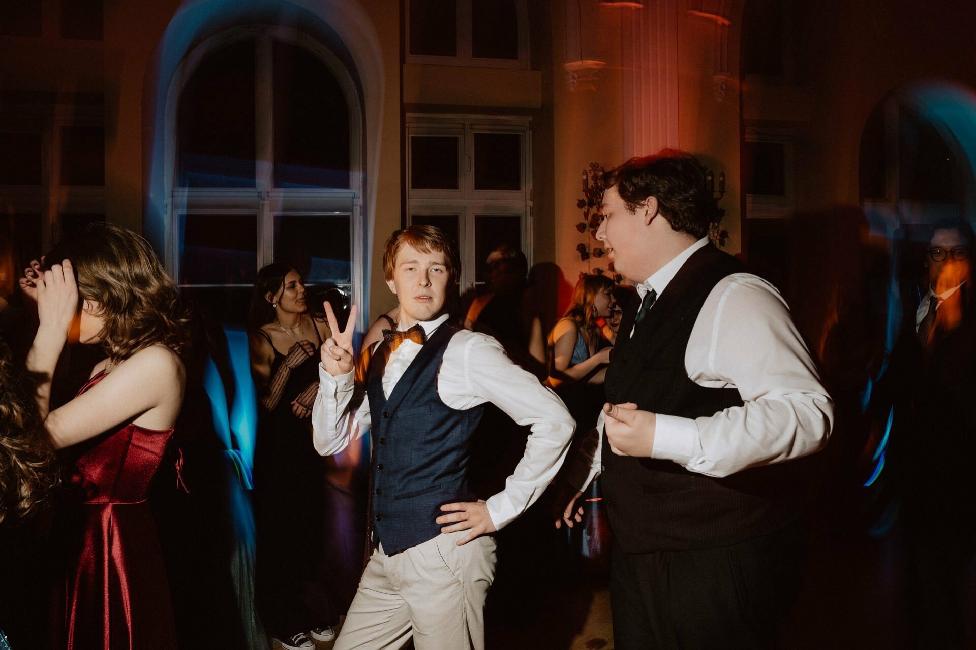 Young adults dressed formally dance in a dimly lit room, capturing the essence of "The Prom Report." The person in the center gives the peace sign, wearing a blue vest and orange tie, while another person on the right improves his bow tie. Others dance in the background under colorful lighting.  