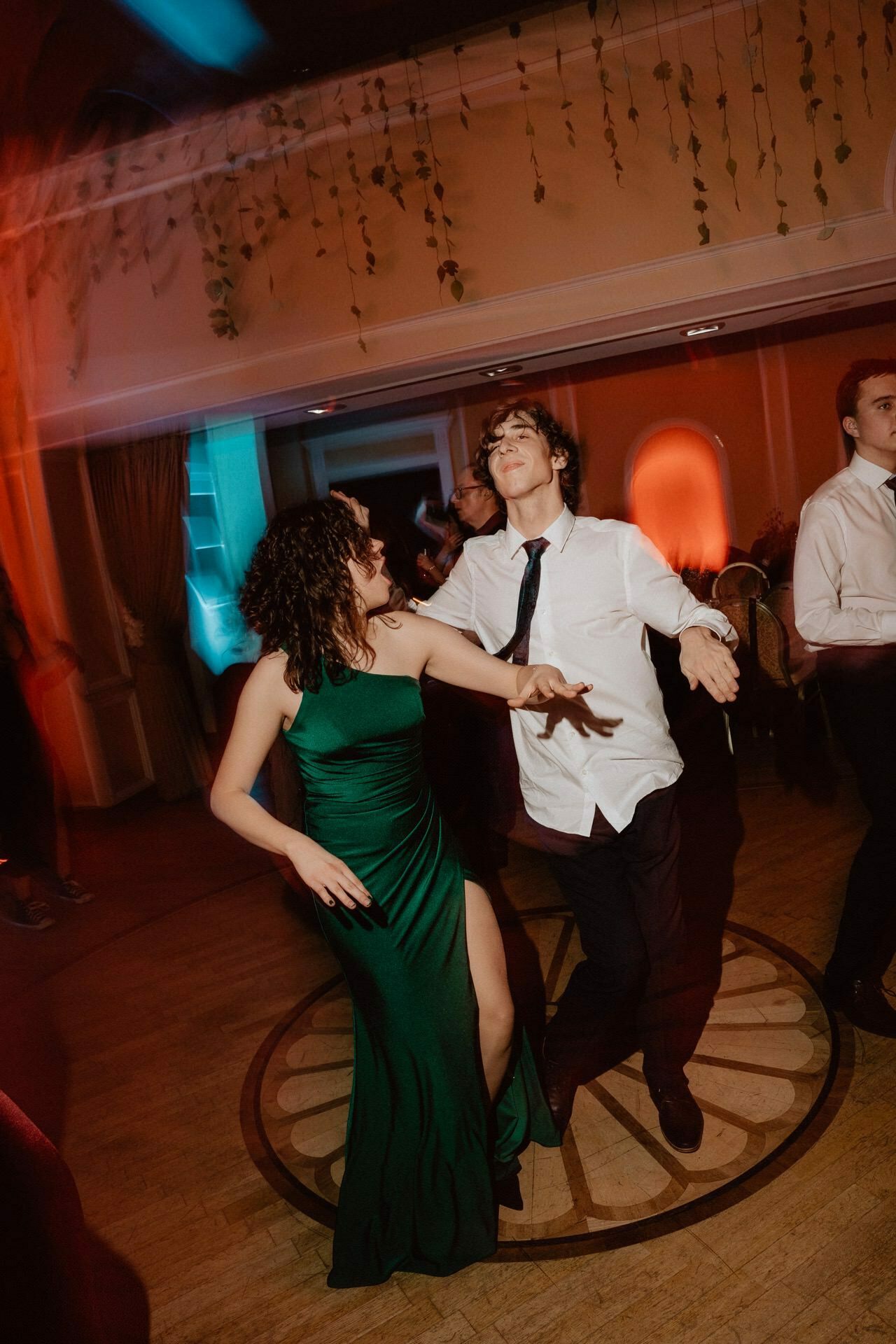 People dance in a warmly lit room decorated with hanging vines. A woman in a long green dress dances energetically with a man in a white shirt and tie. In the background you can see other people dancing, creating a lively and joyful atmosphere - a charming reportage of a prom.  