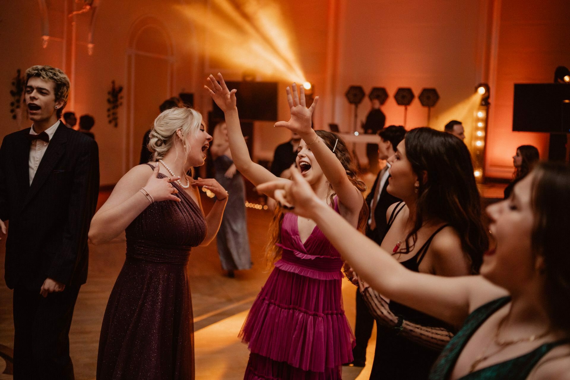 A vibrant scene unfolds during a party held in a room with warm, dramatic lighting. The reportage from the prom captures the joy as a group of people exuberantly dance and sing. In the foreground, two women in bright purple and pink ruffled dresses enthusiastically gesture, while others add to the festive atmosphere in the background.  