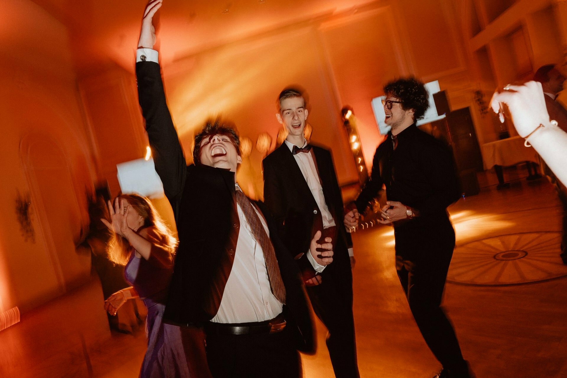 A group of formally dressed people joyfully dance in a warmly lit room during a report from a prom. One person raises his hand in the air, another smiles, and there is a lively, festive atmosphere with fuzzy movement that adds energy to the scene. 